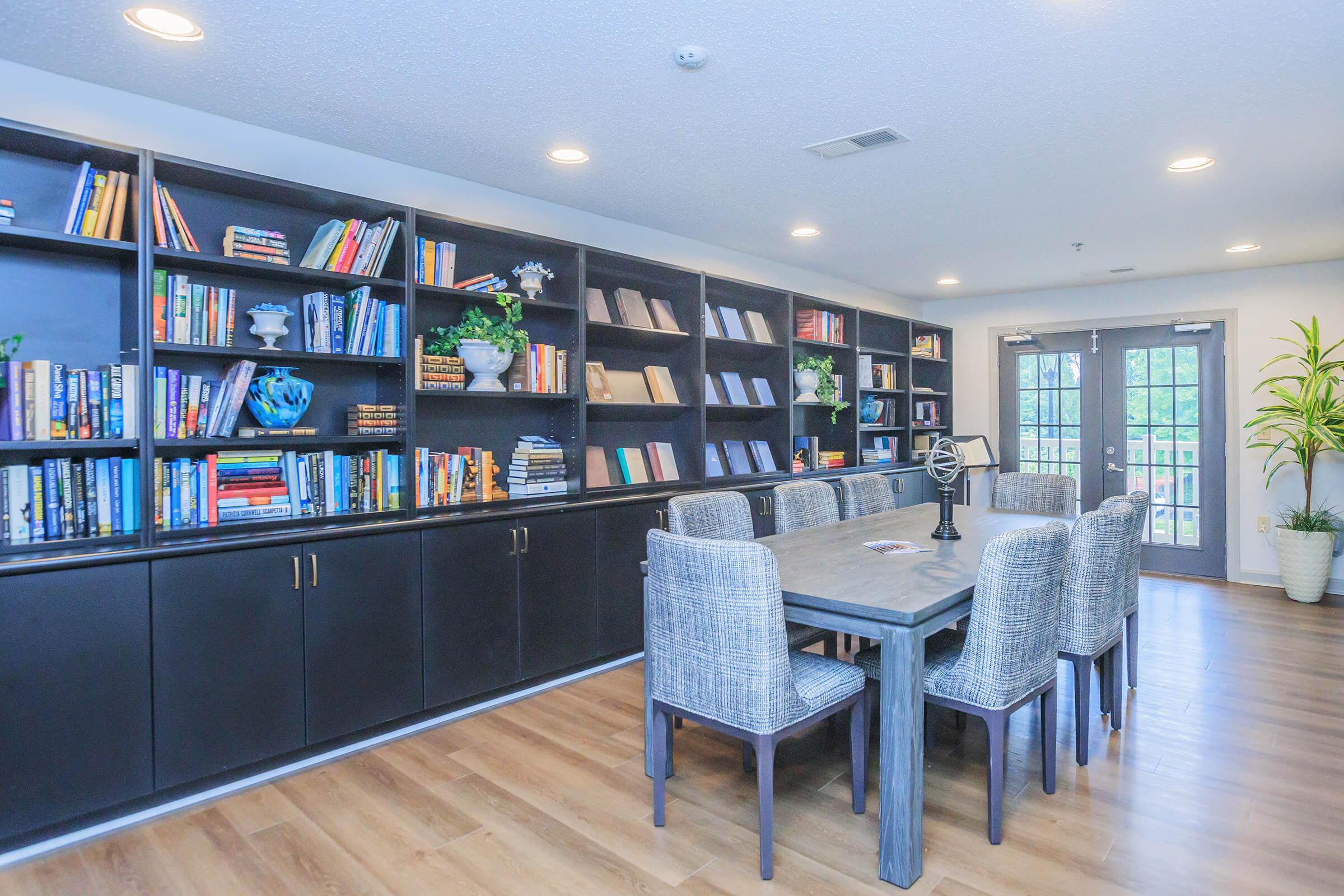 A cozy reading room featuring a large bookshelf filled with books, a wooden dining table surrounded by woven chairs, and a decorative globe. Large windows provide natural light, and indoor plants add a touch of greenery to the space.