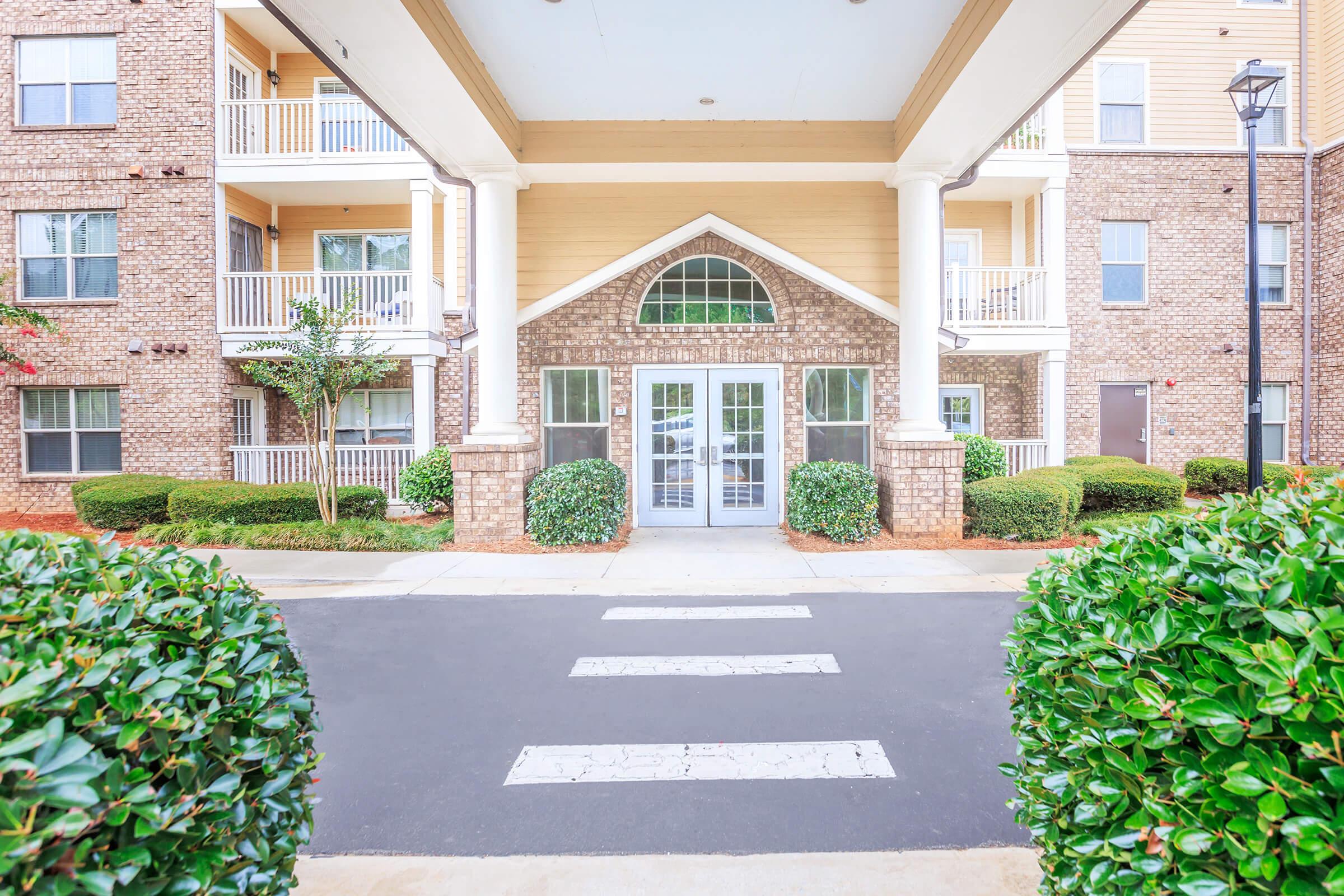 View of an apartment complex entrance featuring double glass doors framed by white columns. The building features a blend of brick and wood siding, with lush green bushes and trees around the entrance. A crosswalk leads toward the entrance, enhancing accessibility. Natural light brightens the area.