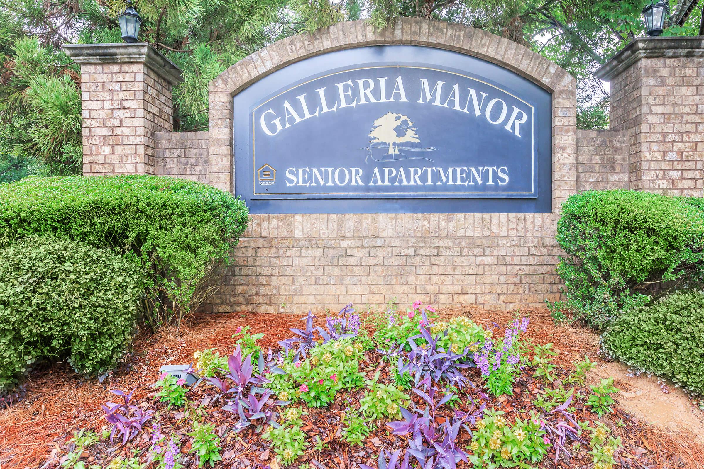 Sign for Galleria Manor Senior Apartments, featuring a brick structure with manicured greenery and colorful flowerbeds. The sign has a dark background with white text, prominently displaying the name and purpose of the complex.