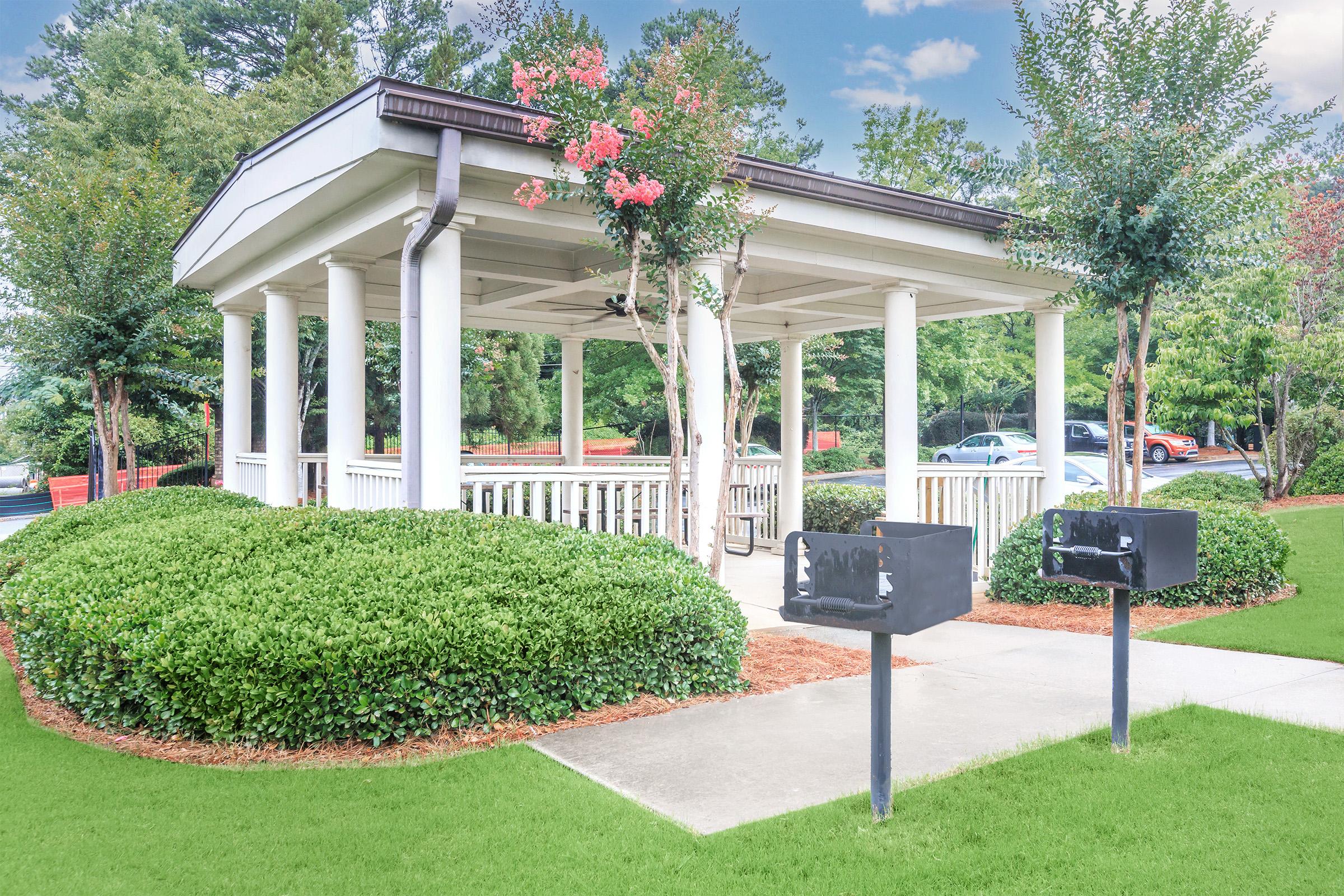 A small gazebo surrounded by greenery, featuring several white columns and colorful flowering plants. Two black mailboxes stand in front on a concrete path, with manicured bushes and grass in the foreground. Trees and vehicles are visible in the background under a clear sky.