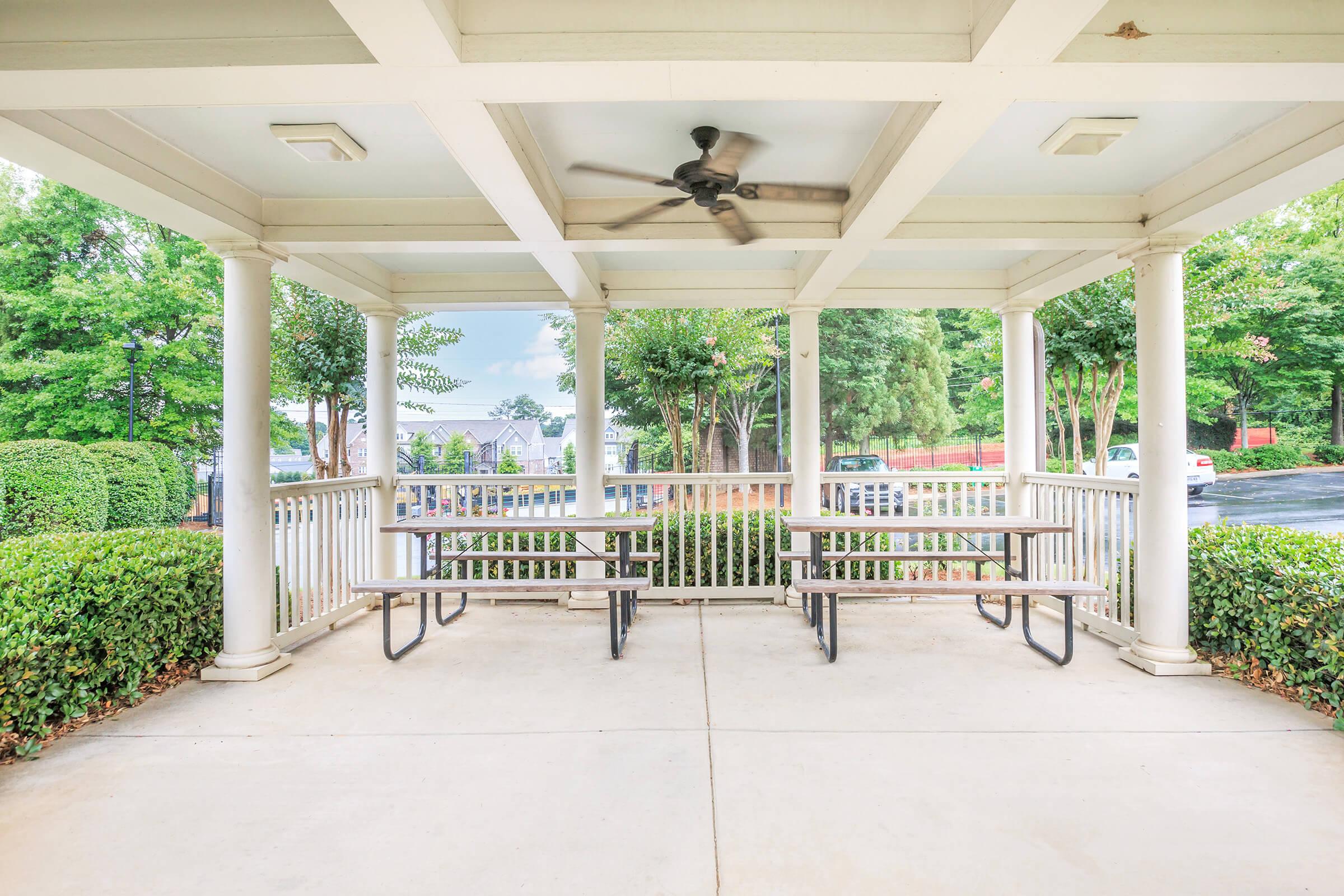 A covered outdoor seating area featuring several picnic tables surrounded by greenery. The space is well-lit with a ceiling fan and has columns supporting the structure. In the background, there are trees and a glimpse of parked cars. The atmosphere is inviting and suitable for gatherings.