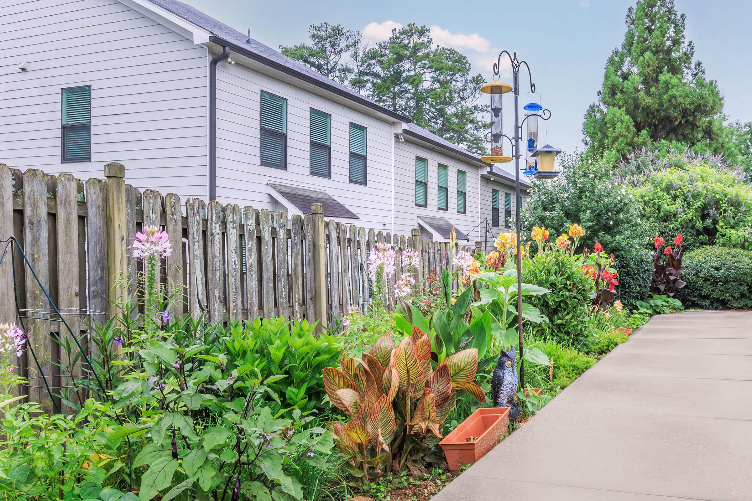 A well-maintained garden featuring a variety of colorful flowers and lush green plants, bordered by a wooden fence. In the background, two white houses with green shutters are visible, along with bird feeders and a clear blue sky. A concrete pathway runs alongside the garden, adding to the serene outdoor atmosphere.