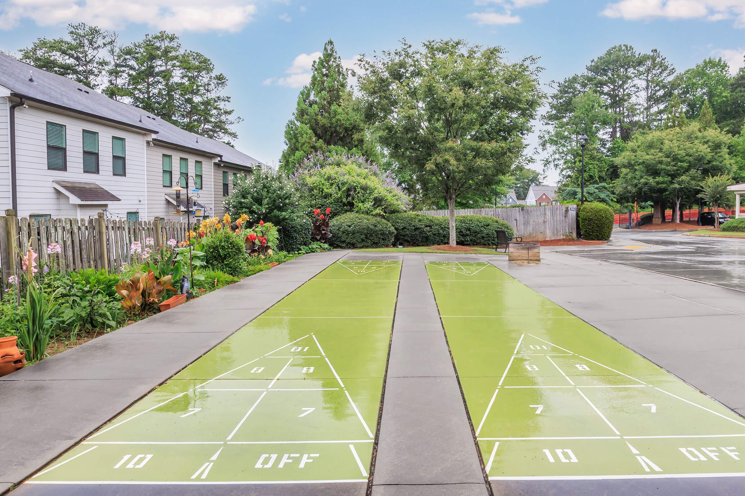 Two shuffleboard courts in a residential area, set on a smooth, green surface with white lines marking the scoring sections. Surrounding the courts are colorful flowerbeds and well-maintained foliage. In the background, there's a fence and several houses under a partly cloudy sky.