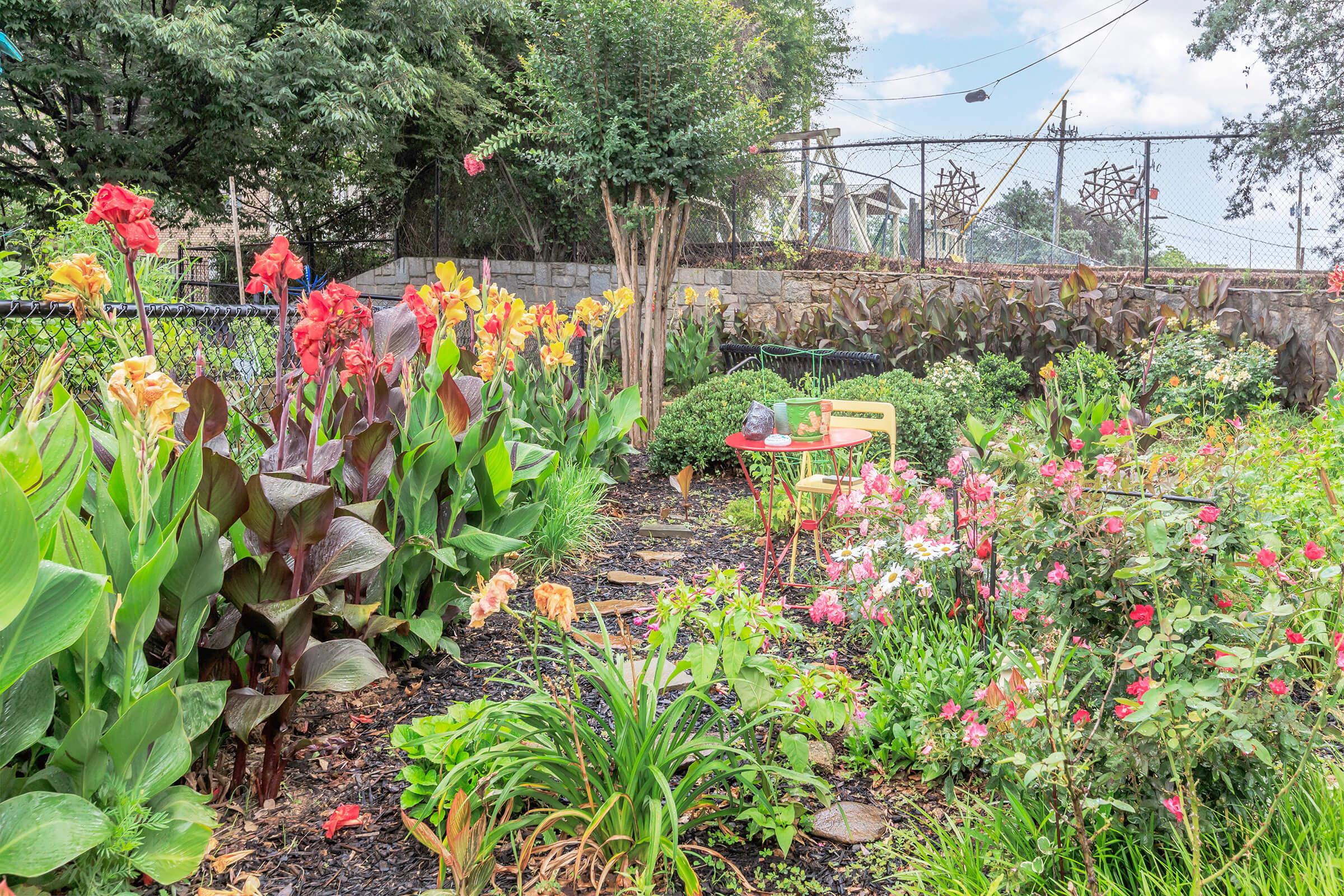 A vibrant garden filled with colorful flowers, including red and yellow canna lilies, pink begonias, and greenery. In the center, a wooden chair sits amidst the blossoms, inviting relaxation. The background features a fence and trees, with a blue sky peeking through scattered clouds.