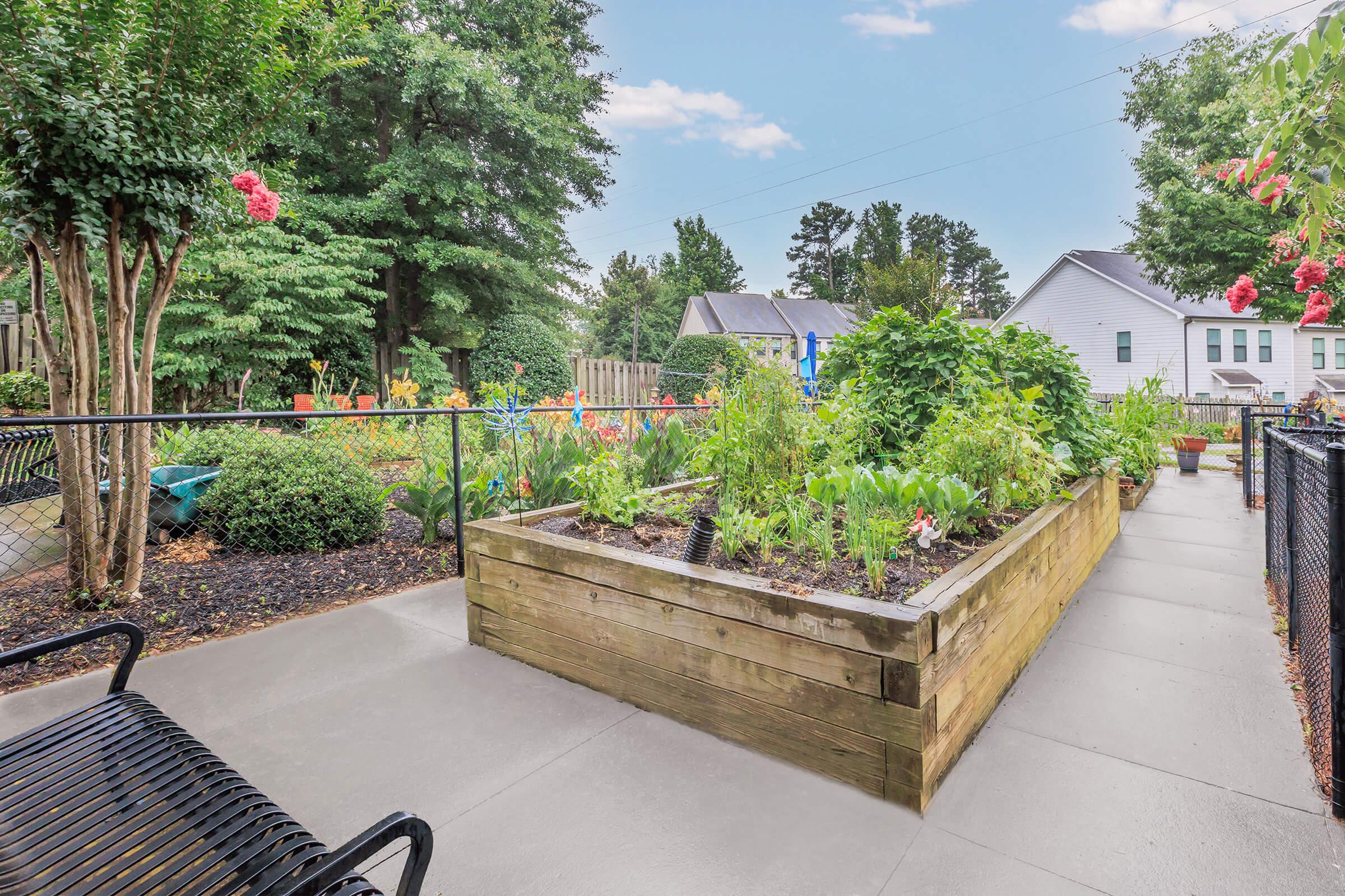 A peaceful garden scene featuring a wooden raised bed filled with various plants and vegetables, surrounded by green foliage and vibrant flowers. A concrete pathway runs alongside the garden, with black benches for sitting, creating an inviting outdoor space. Calm and serene atmosphere.