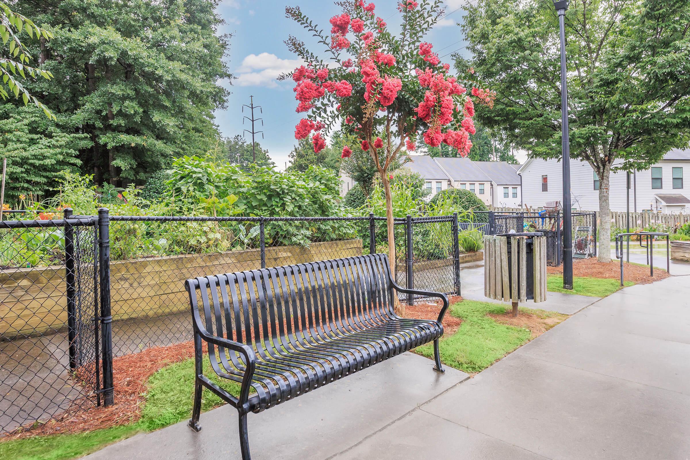 A park scene featuring a black bench in the foreground, lush greenery including flowering trees, and a fenced garden area. In the background, there are residential buildings and a trash bin. The sky is partly cloudy, indicating a pleasant day.