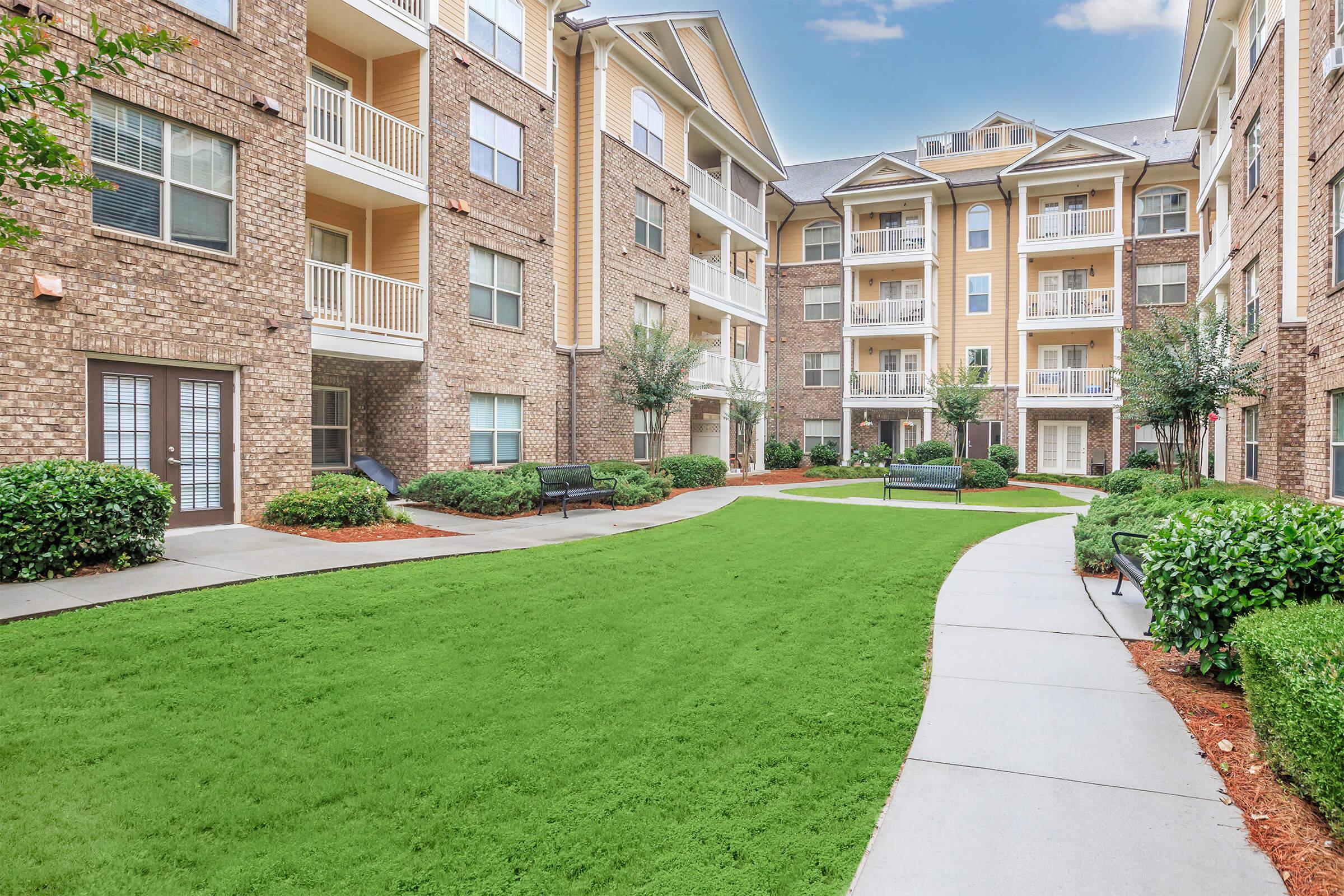 A landscaped courtyard in an apartment complex featuring manicured green grass, walkways, and several benches. The buildings are multi-story with a mix of brick and siding, and there are small trees and shrubs enhancing the outdoor area. The sky is clear with a few clouds.