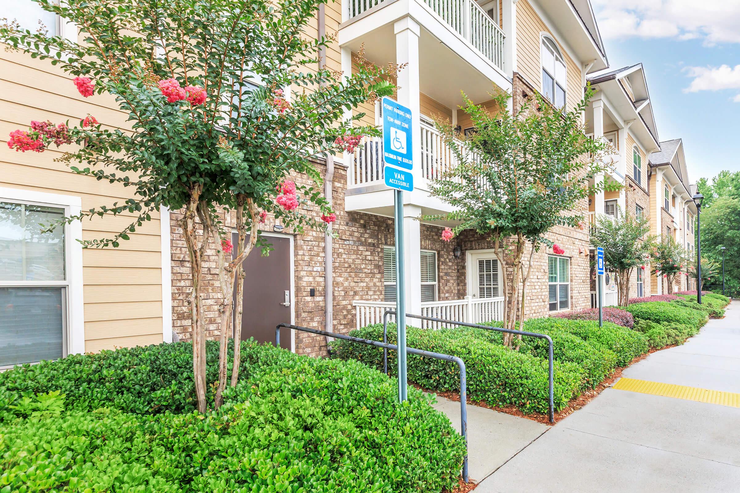 A well-maintained apartment building with brick and beige siding. Lush green landscaping, including shrubs and flowering plants, frames the entrance. A blue sign indicating accessible parking is visible near the pathway, which leads to the building. The sky is clear, adding to the inviting appearance of the property.