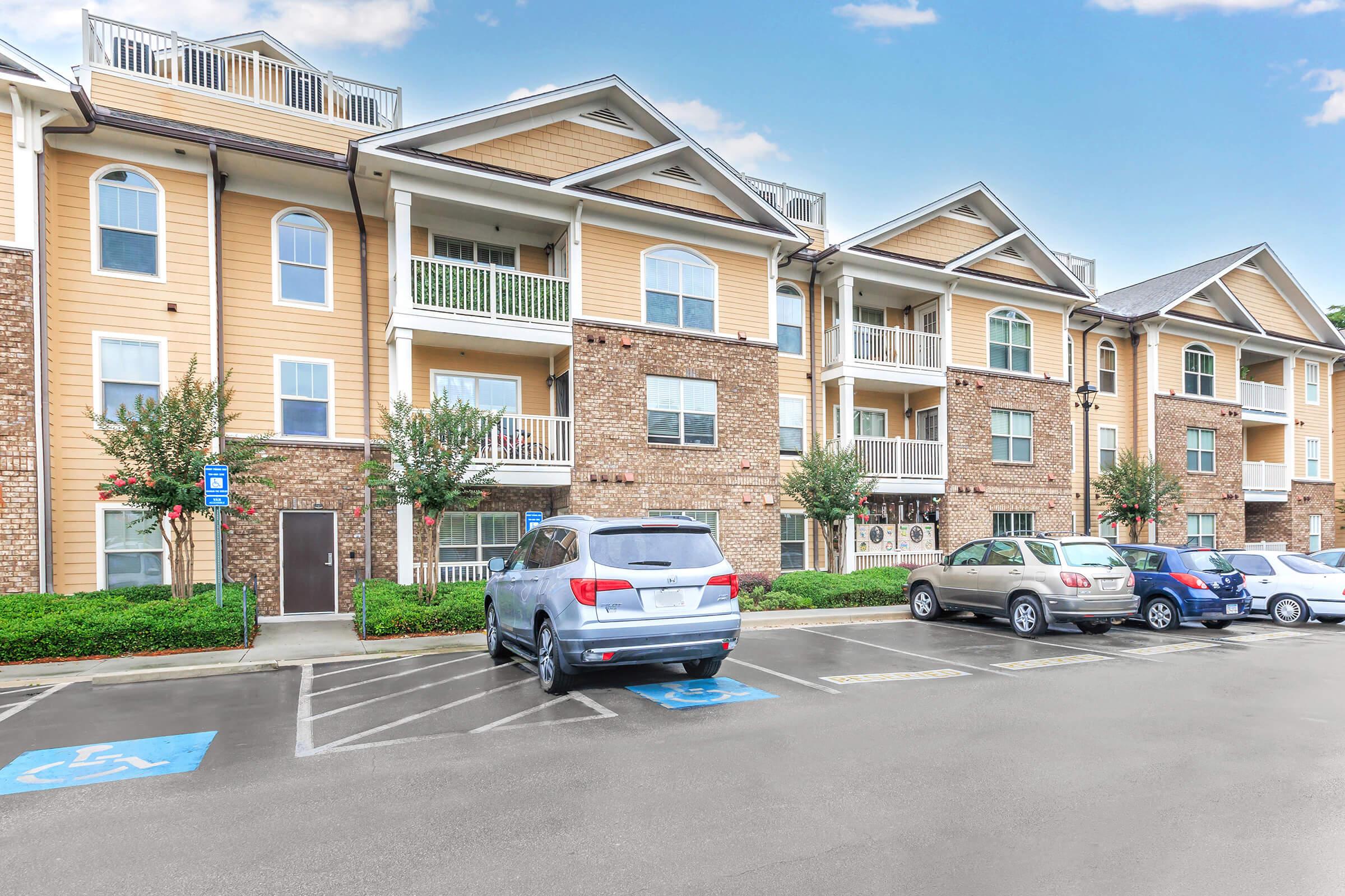 A residential apartment complex featuring multiple floors with a mix of tan and brick exteriors. Balconies are visible on the upper levels. The parking lot shows several cars, including a gray SUV in a handicapped spot, surrounded by landscaped green areas. Clear blue sky in the background.
