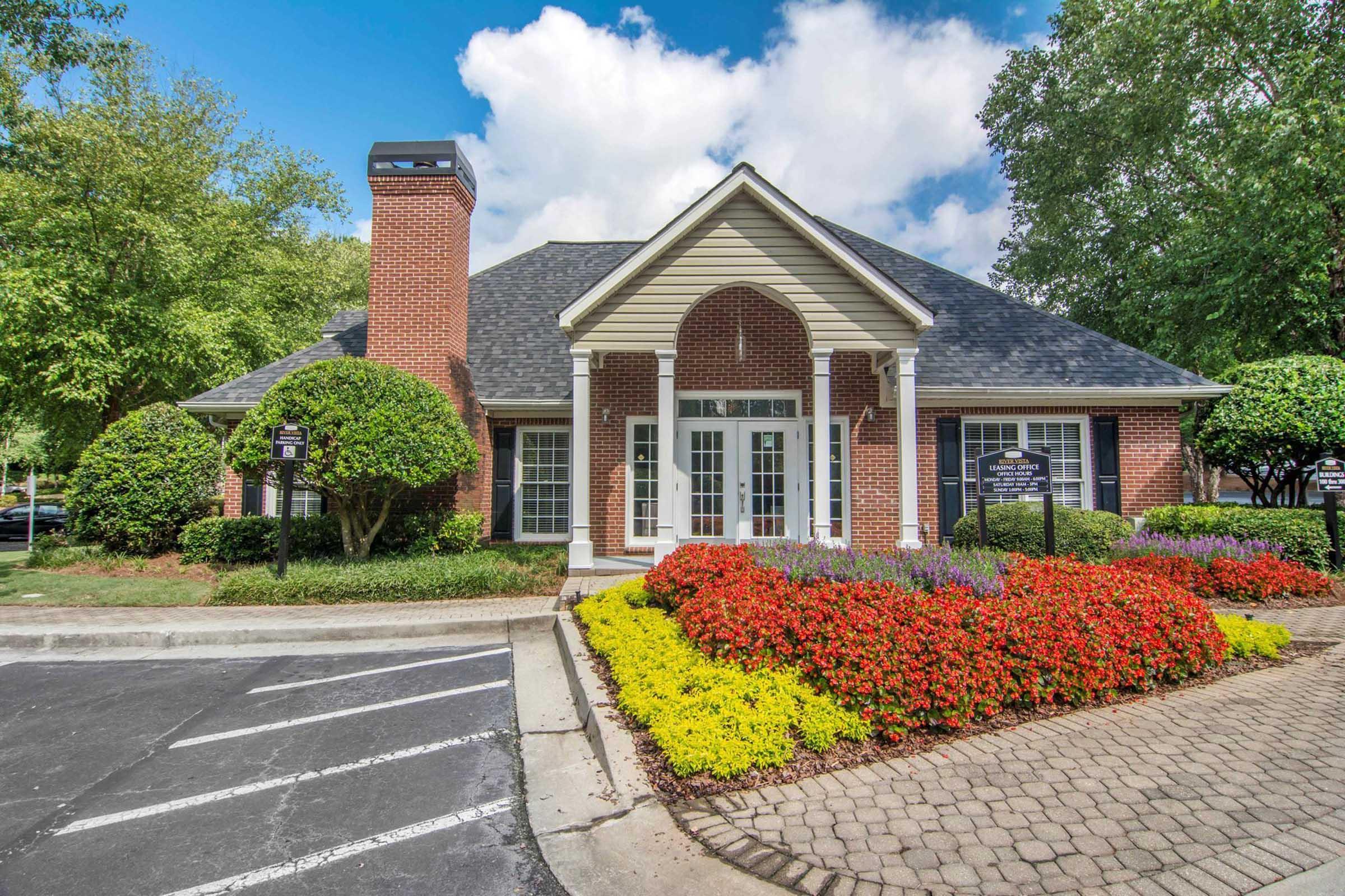 A welcoming brick building with a gabled roof, surrounded by vibrant flower beds featuring red, yellow, and purple blooms. Lush green bushes are neatly trimmed, and a paved pathway leads to the entrance. The sky is partly cloudy, adding a sunny atmosphere to the scene.