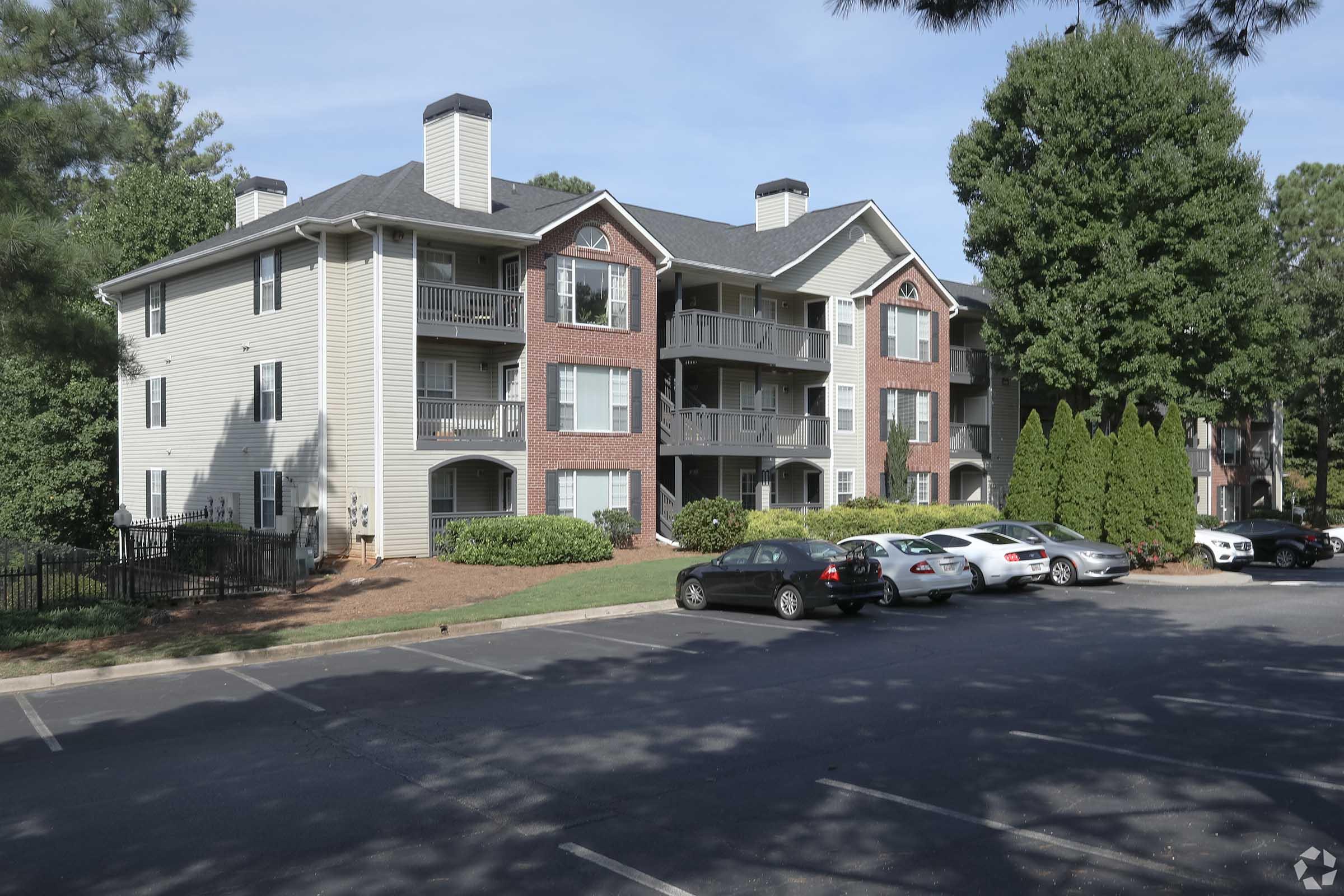 Apartment building with a mix of brick and siding exterior, featuring multiple balconies. Surrounding the building are landscaped areas with shrubs and trees. A parking lot in front shows several parked cars.