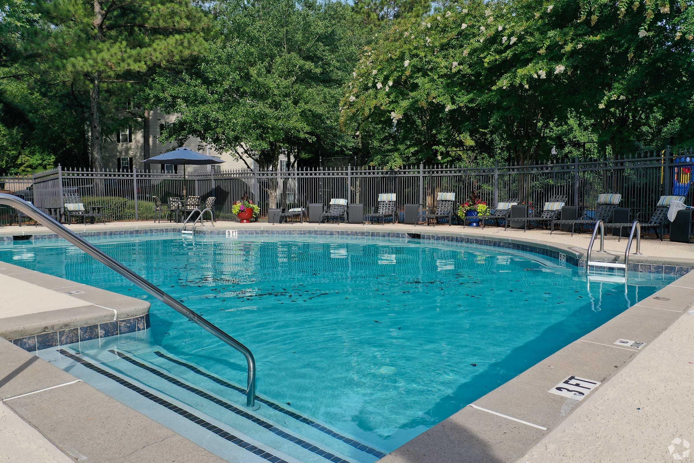 A clear blue swimming pool surrounded by a concrete deck, with steps leading into the water. There are several lounge chairs arranged around the pool and a fence in the background. Trees provide shade, and there are some potted plants nearby. No people are present in the scene.