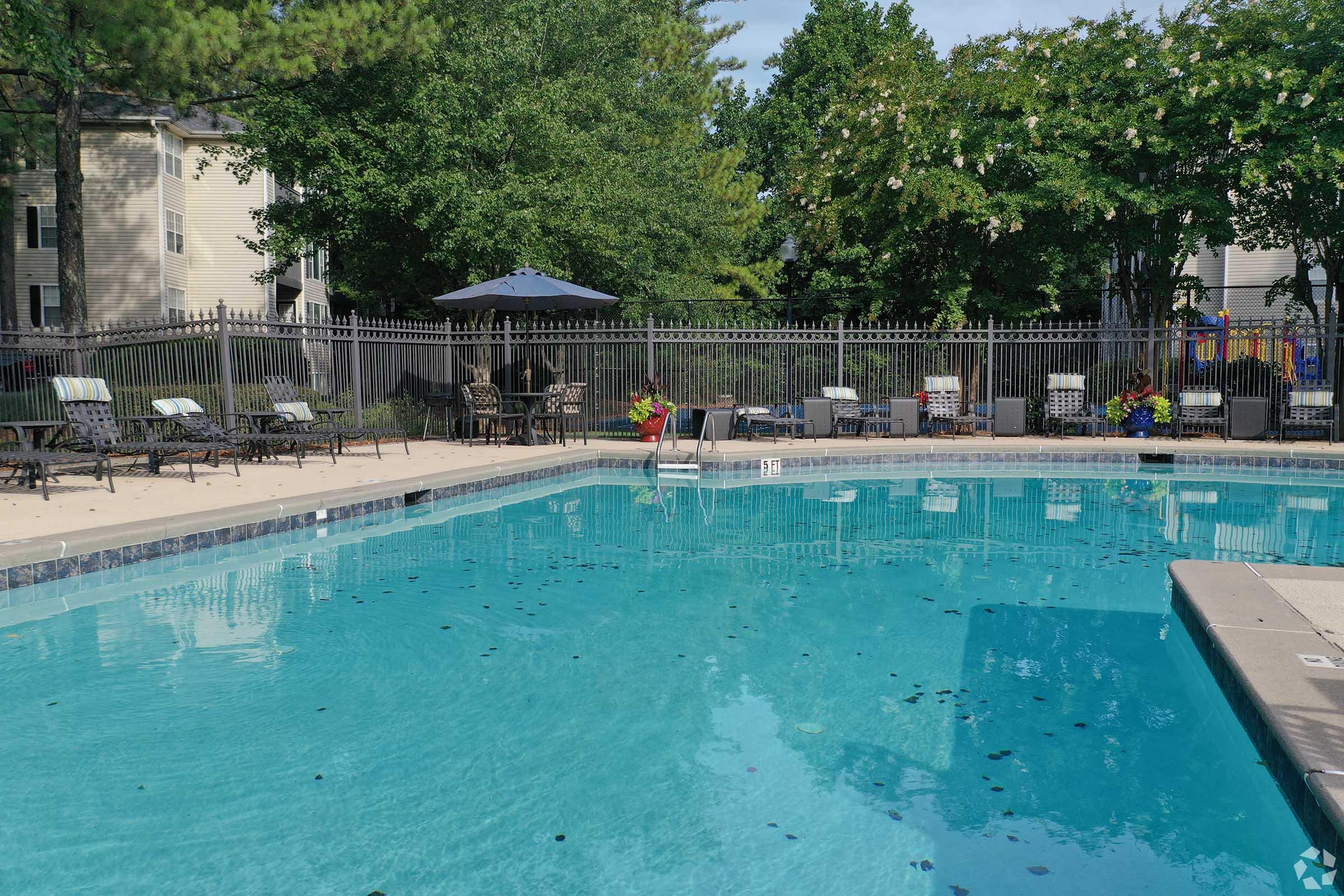A clear blue swimming pool surrounded by a patio area, featuring lounge chairs, umbrellas, and potted plants. A fence encloses the pool, with trees in the background and a playground visible in the distance. The scene is sunny and inviting, suggesting a relaxing outdoor space.