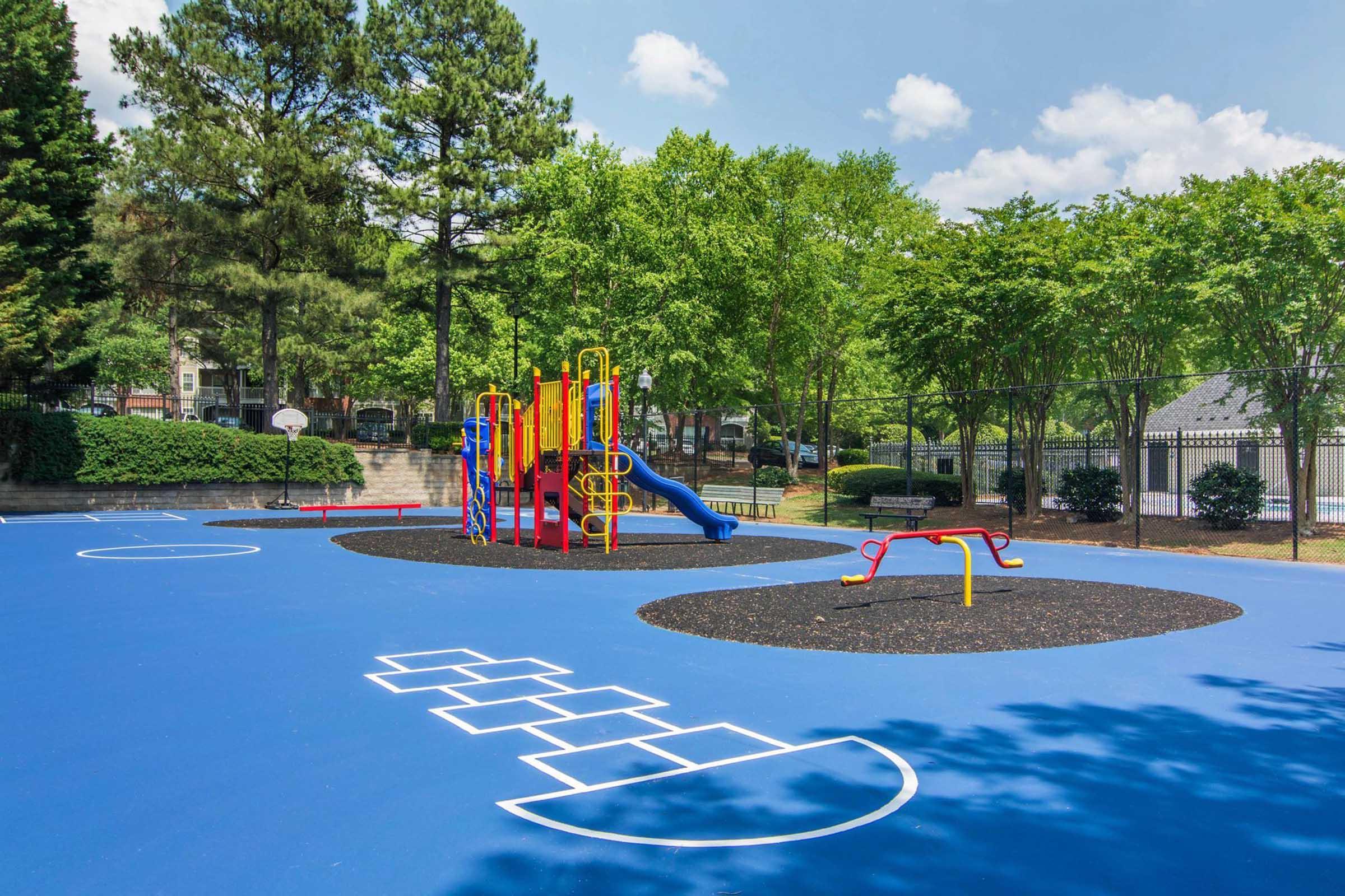 A colorful playground featuring a slide, climbing structure, and swings, set on a vibrant blue rubber surface. Surrounded by green trees and a fence, the area includes a basketball court in the background, providing a fun and safe environment for children to play.