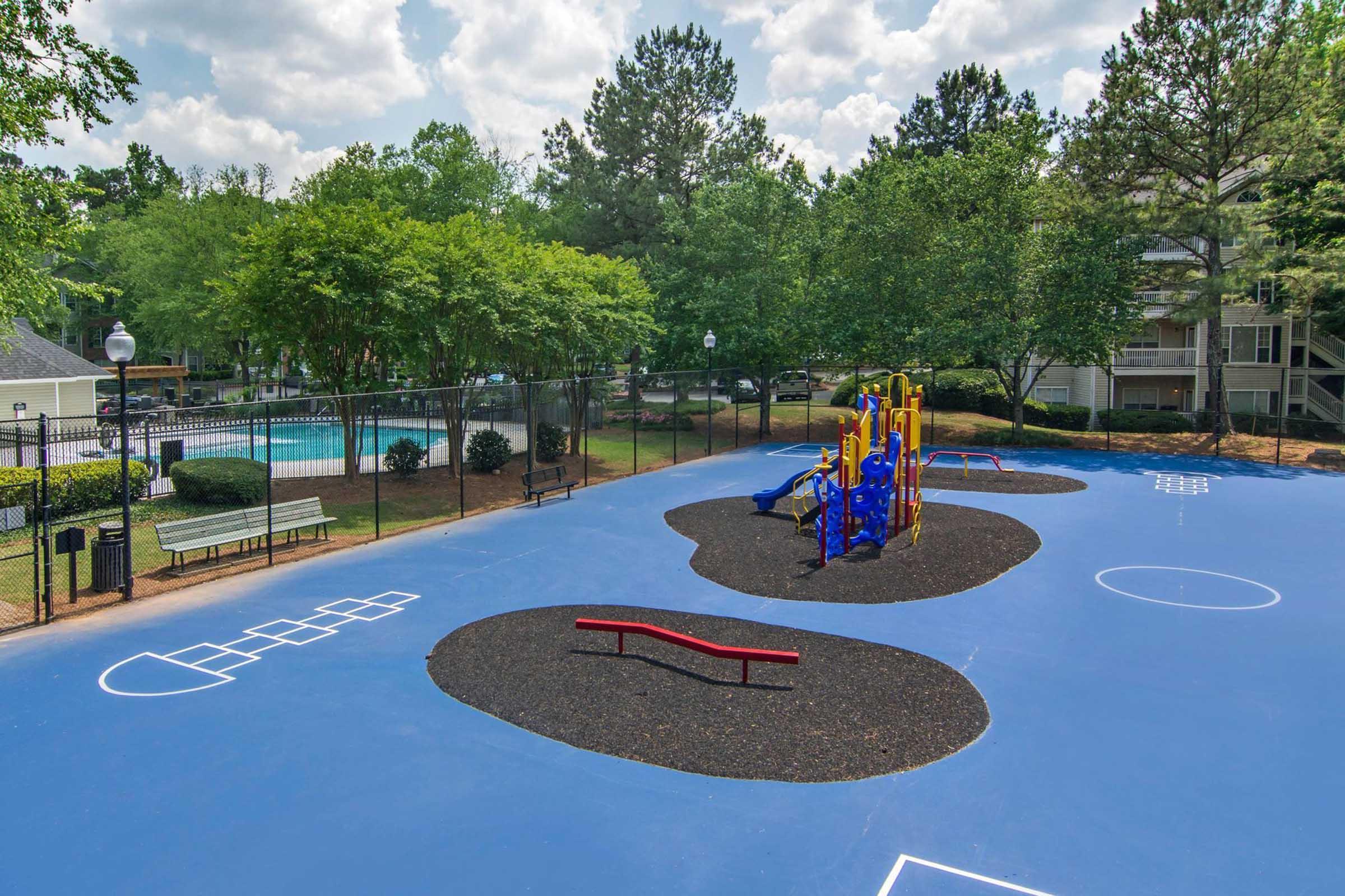 Colorful playground equipment on a blue surface, surrounded by black rubber mulch. The area features various climbing structures, slides, and a balance beam. Nearby, a swimming pool and benches are visible amidst lush green trees. The scene is bright and sunny, conveying a welcoming atmosphere for children.