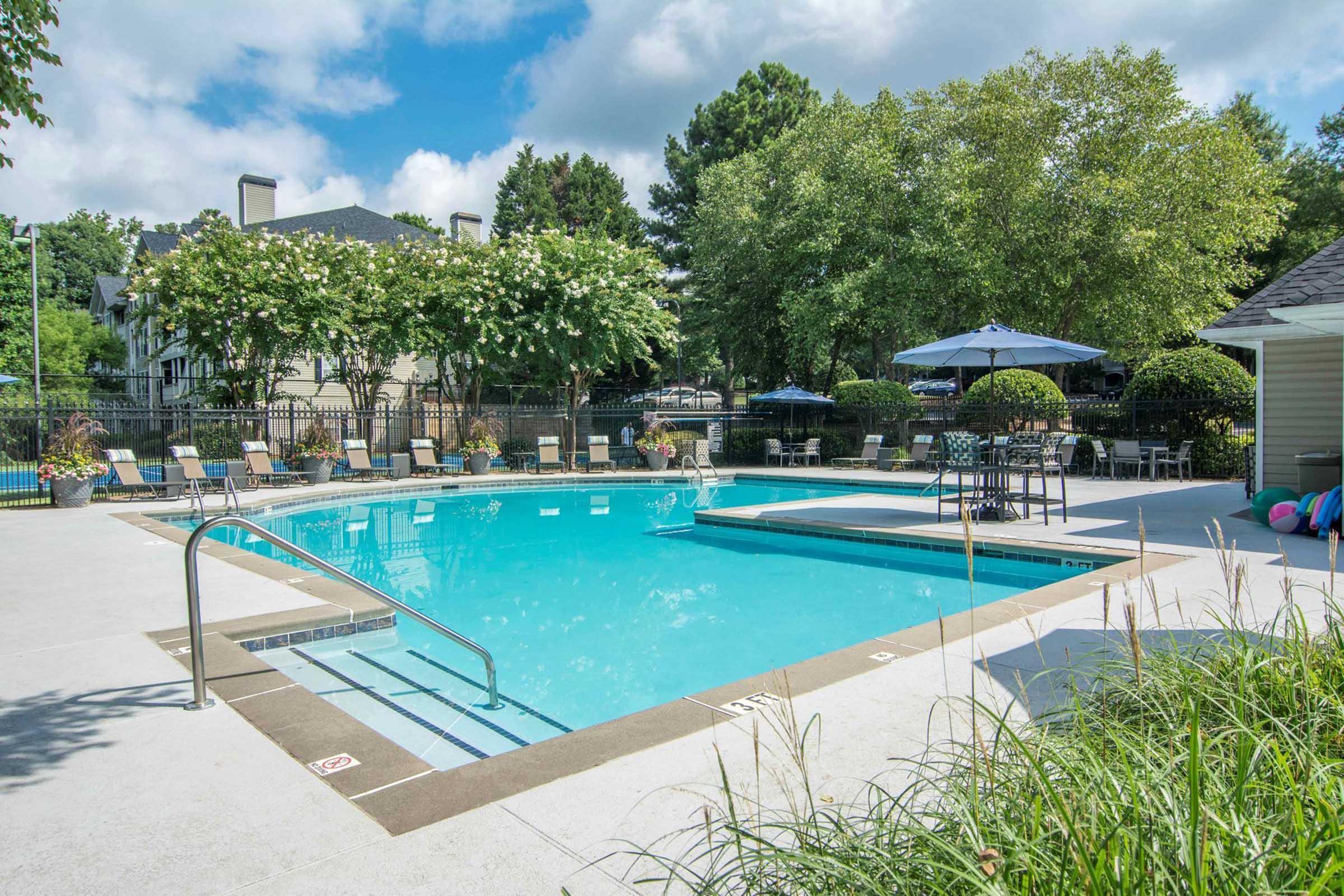 A bright outdoor swimming pool surrounded by lush greenery and lounge chairs. Umbrellas provide shade around the pool area, which is fenced for safety. In the background, there are residential buildings and colorful flowers. The sky is partly cloudy, creating a serene atmosphere.