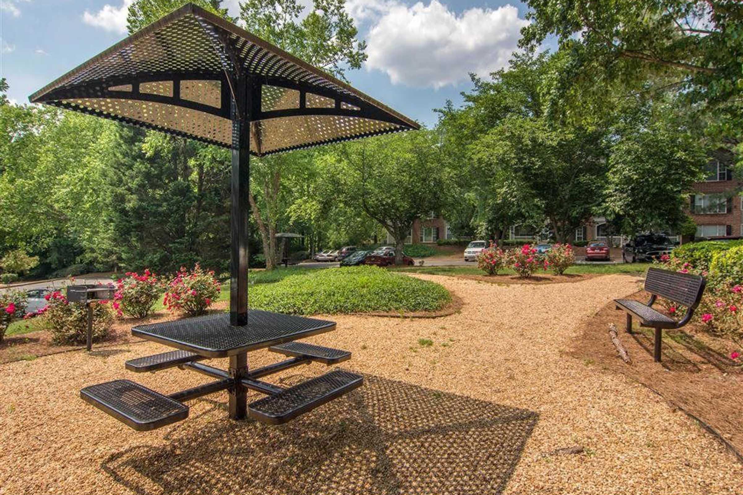 A park area featuring a black metal picnic table with an umbrella, surrounded by lush greenery and colorful flower beds. A gravel path leads to the table, with benches nearby and parked cars visible in the background under a partly cloudy sky.
