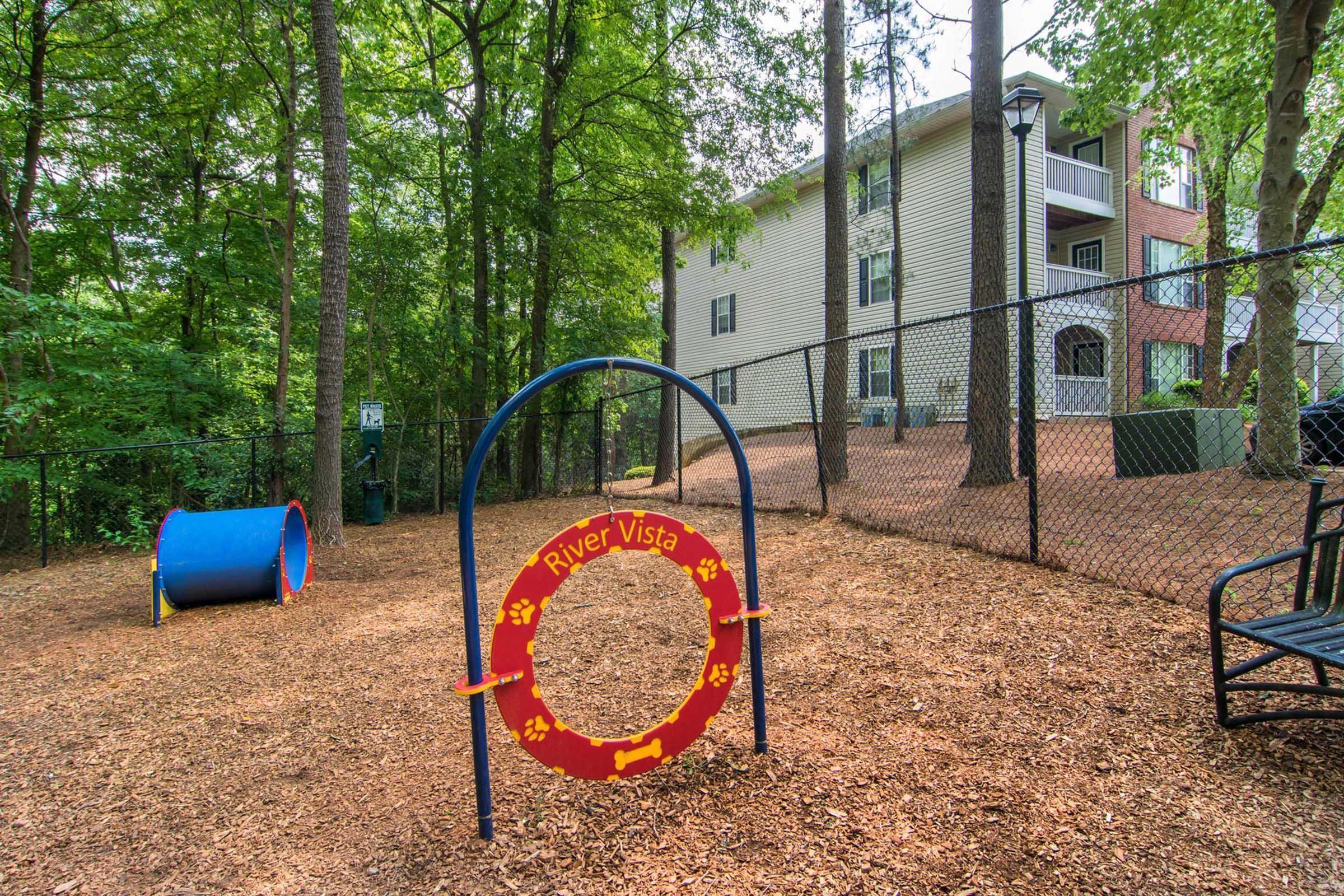 A playground area featuring a red play tunnel and a circular hoop with "River Vista" written on it, surrounded by mulch and trees. In the background, there's an apartment building with a fenced yard, providing a natural and inviting space for children to play.