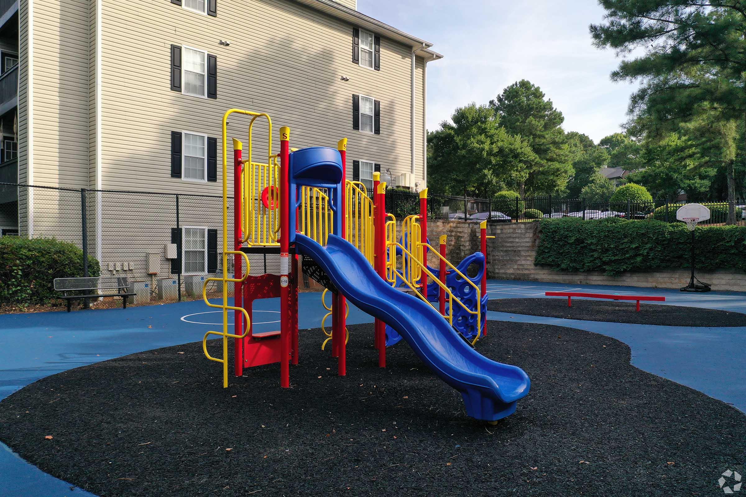 A colorful children's playground featuring a blue slide, climbing structure, and red and yellow play equipment. The area has a black rubber surface and is surrounded by greenery and a residential building in the background. A basketball court is partially visible on the right.