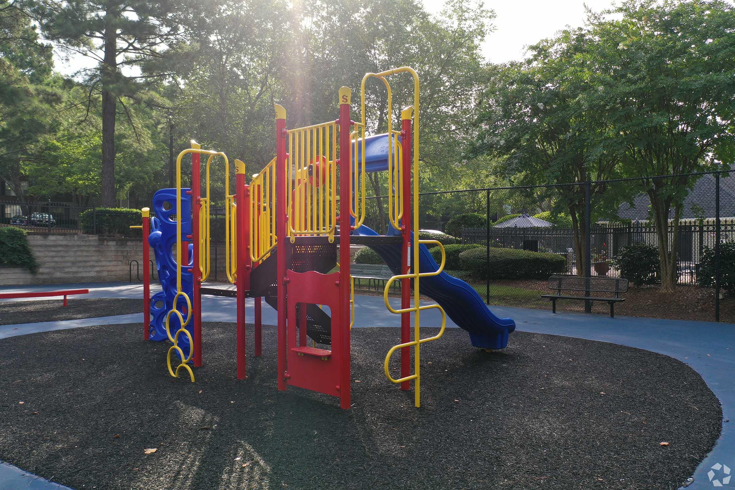 Colorful children's playground with slides and climbing structures, set in a park surrounded by trees. The play area features blue rubber surfacing and gravel, with benches nearby. Sunlight filters through the trees, creating a bright and inviting atmosphere for play.