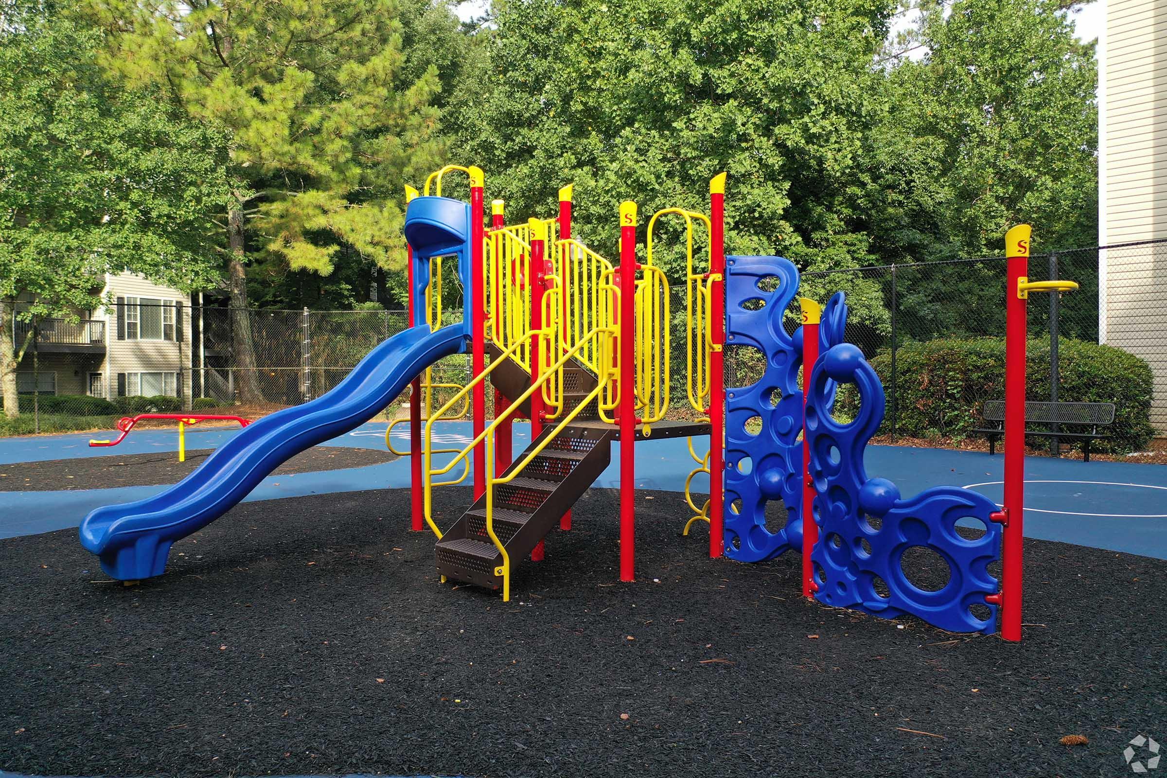 Colorful playground equipment featuring a blue slide, yellow and red climbing structures, and textured climbing walls. The play area is surrounded by a soft black surface and green trees in the background, creating a vibrant outdoor space for children to enjoy.