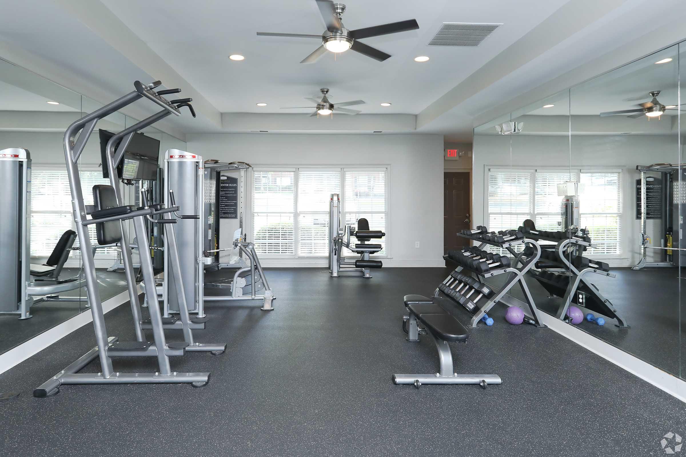 A modern gym interior featuring various exercise equipment, including weight machines and a bench press. The room is well-lit with large windows and ceiling fans, and there are mirrors on one wall. The flooring is dark and rubberized, providing a suitable surface for workouts.