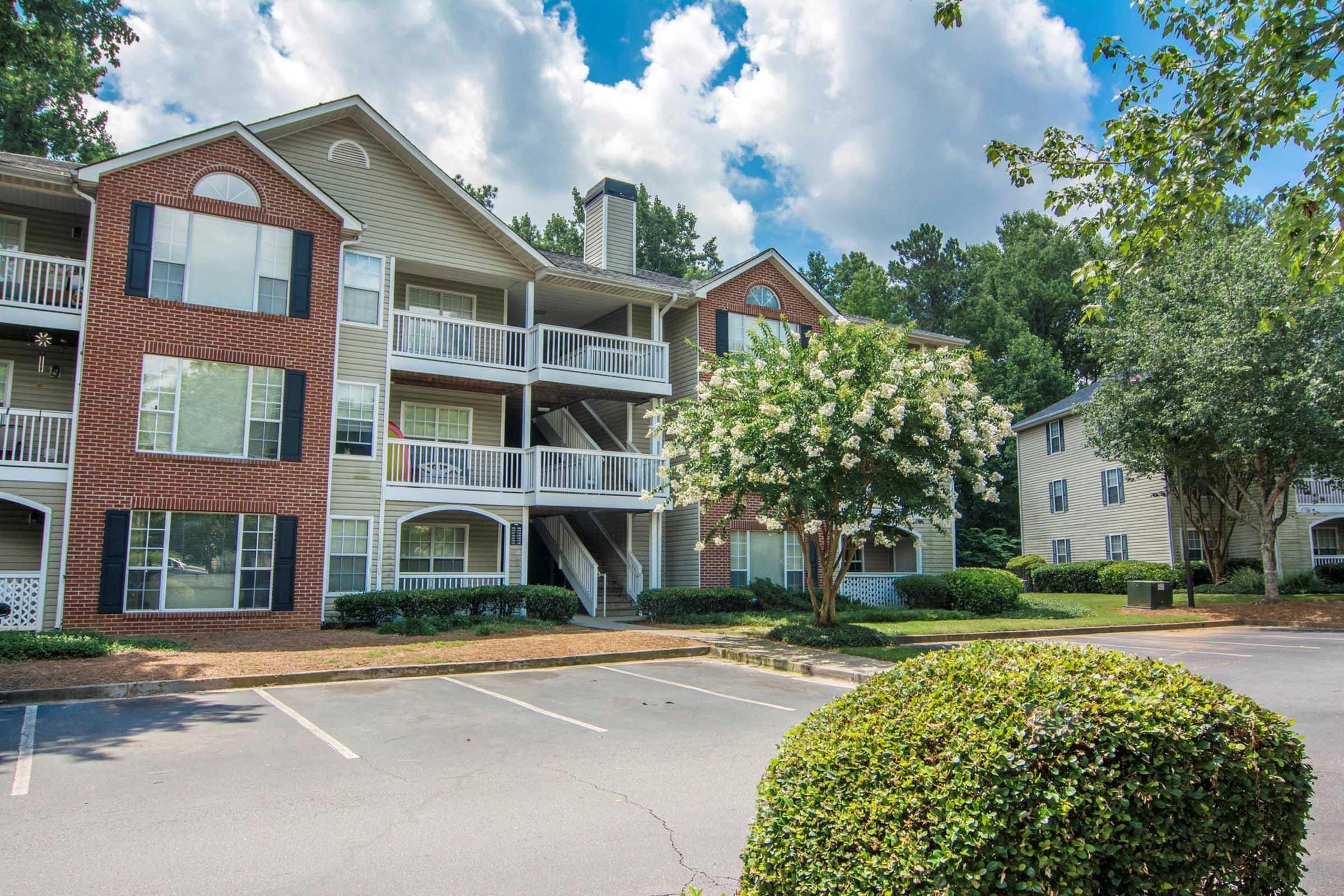 A residential apartment complex featuring three levels with brick and siding accents. The building is surrounded by green lawns, trees, and parking spaces. The sky is partly cloudy, creating a bright atmosphere.