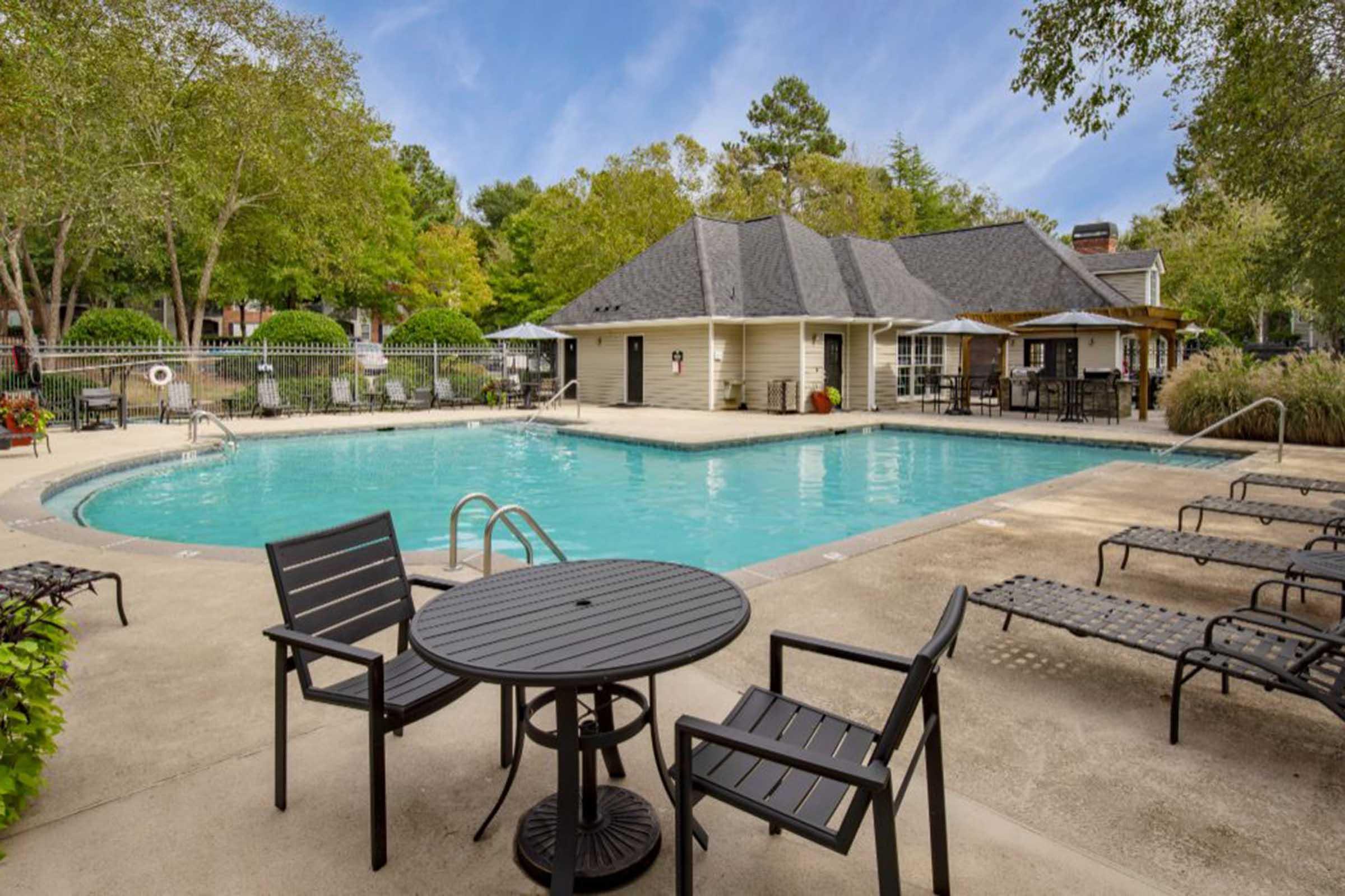 A serene pool area featuring a large swimming pool surrounded by lush greenery. In the foreground, there are black patio tables and chairs, with lounge chairs along the poolside. A clubhouse with a sloped roof is visible in the background, and the sky is clear with a few clouds.