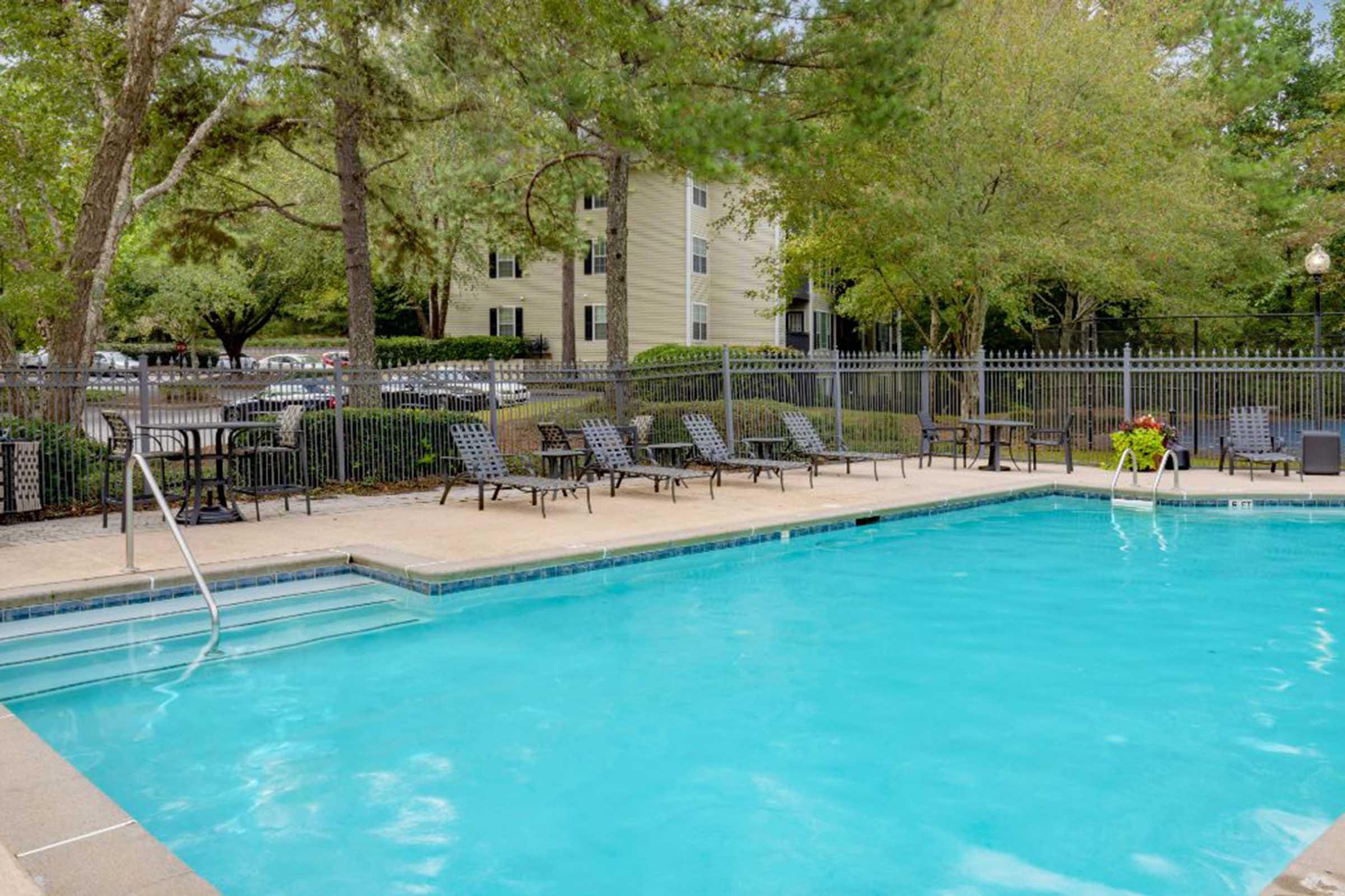 A refreshing swimming pool surrounded by lounge chairs and trees. The pool area features a clean, blue water surface and is enclosed by a fence. In the background, there are apartment buildings and a parking area, creating a relaxing outdoor environment.