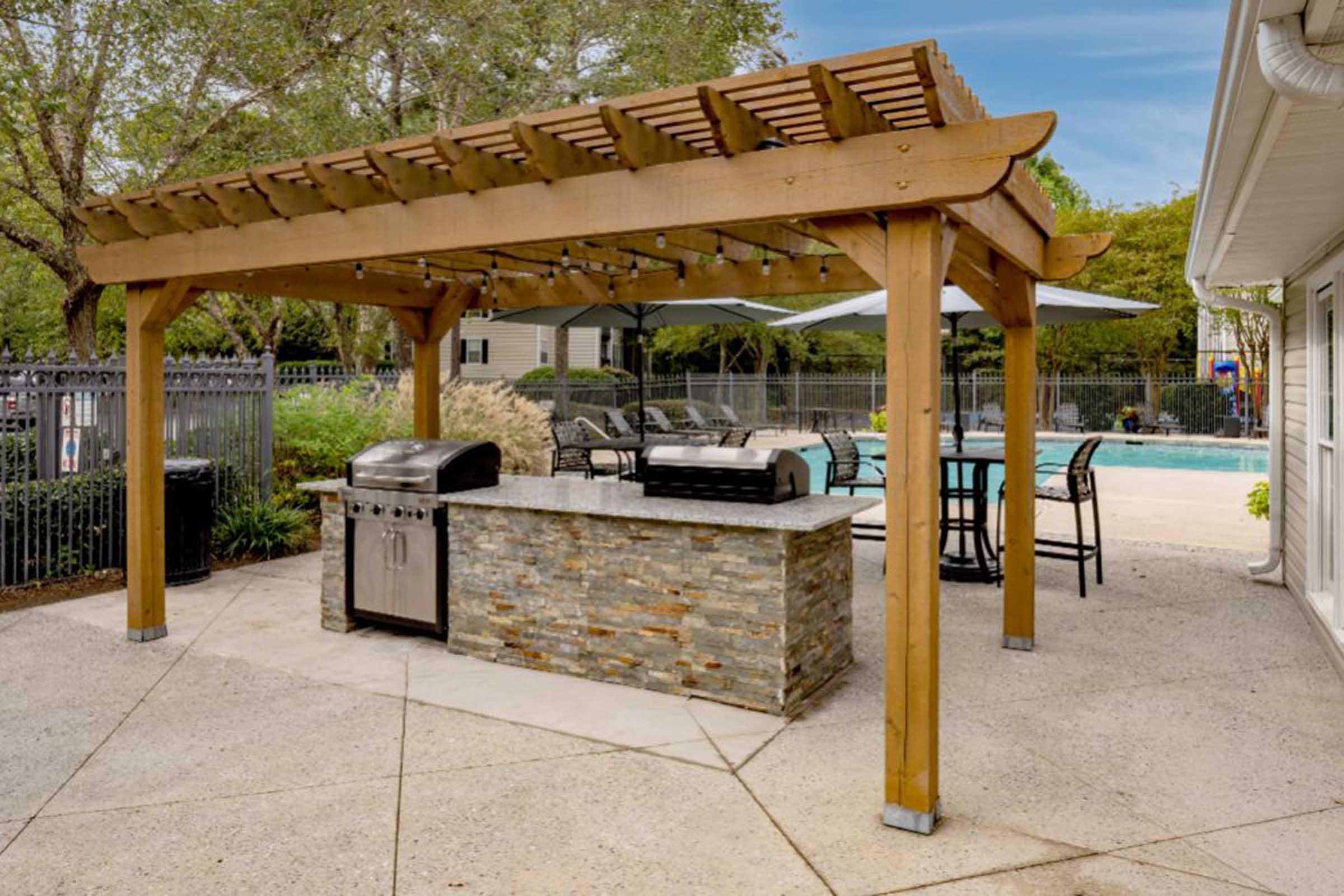 A backyard patio featuring a wooden pergola with a stone grill area. There are two gas grills and several outdoor dining chairs around a table. In the background, a swimming pool is visible, surrounded by lounge chairs and greenery. The area is well-lit and inviting for outdoor gatherings.