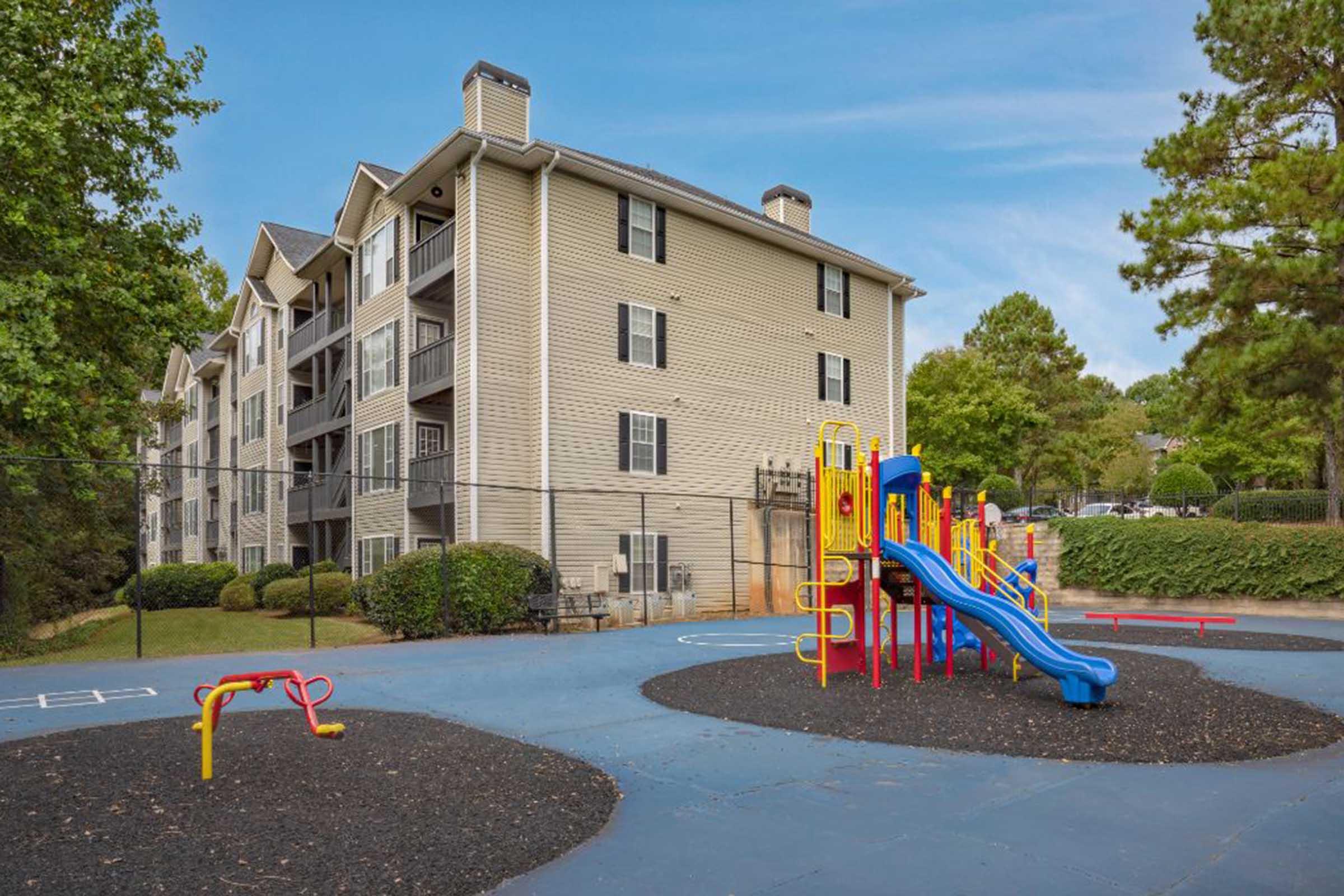 A bright playground featuring colorful slides and swings, surrounded by a black rubber surface. In the background, a three-story apartment building with multiple balconies and green landscaping. A blue sky with a few scattered clouds completes the scene.