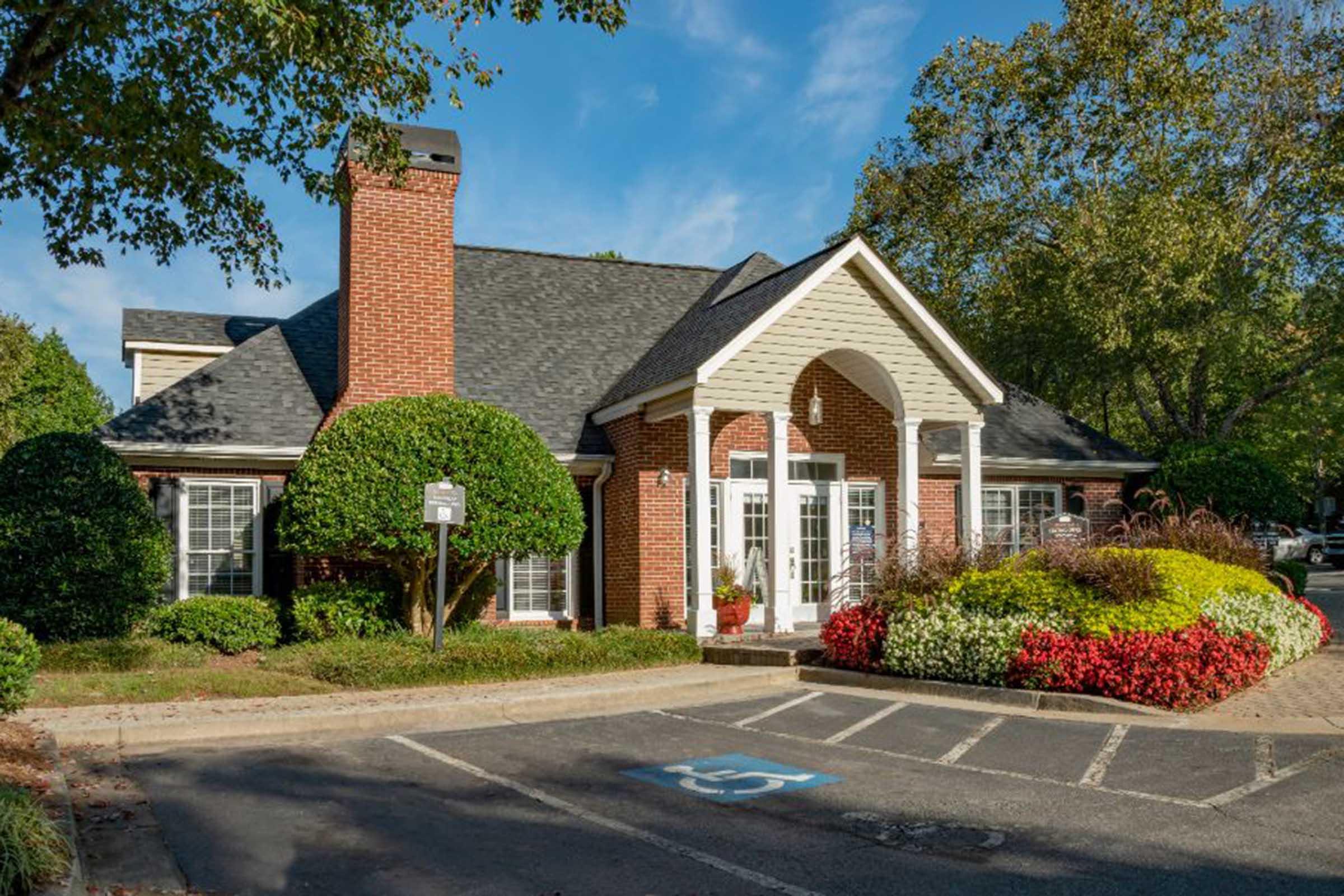 A well-maintained brick building with a peaked roof and white columns at the entrance. Lush green bushes and colorful flower beds frame the front of the building, while a parking lot with a handicap space is visible in the foreground. Clear blue sky and trees provide a serene backdrop.