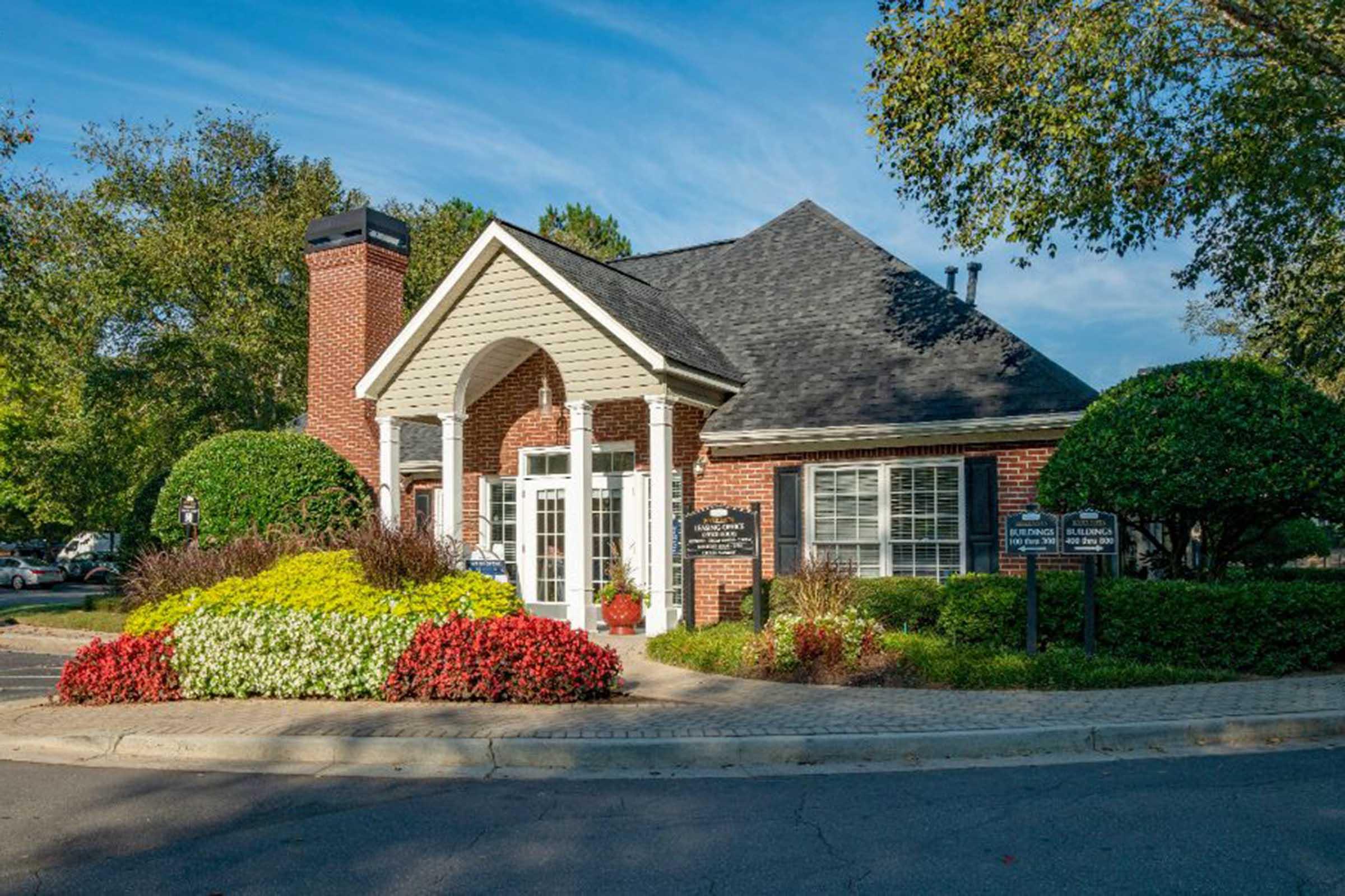 A brick building with a peaked roof and white columns stands surrounded by well-maintained landscaping featuring colorful flower beds and shrubs. The entrance has glass doors, and there are signs nearby indicating the location. Clear blue sky and trees are visible in the background.