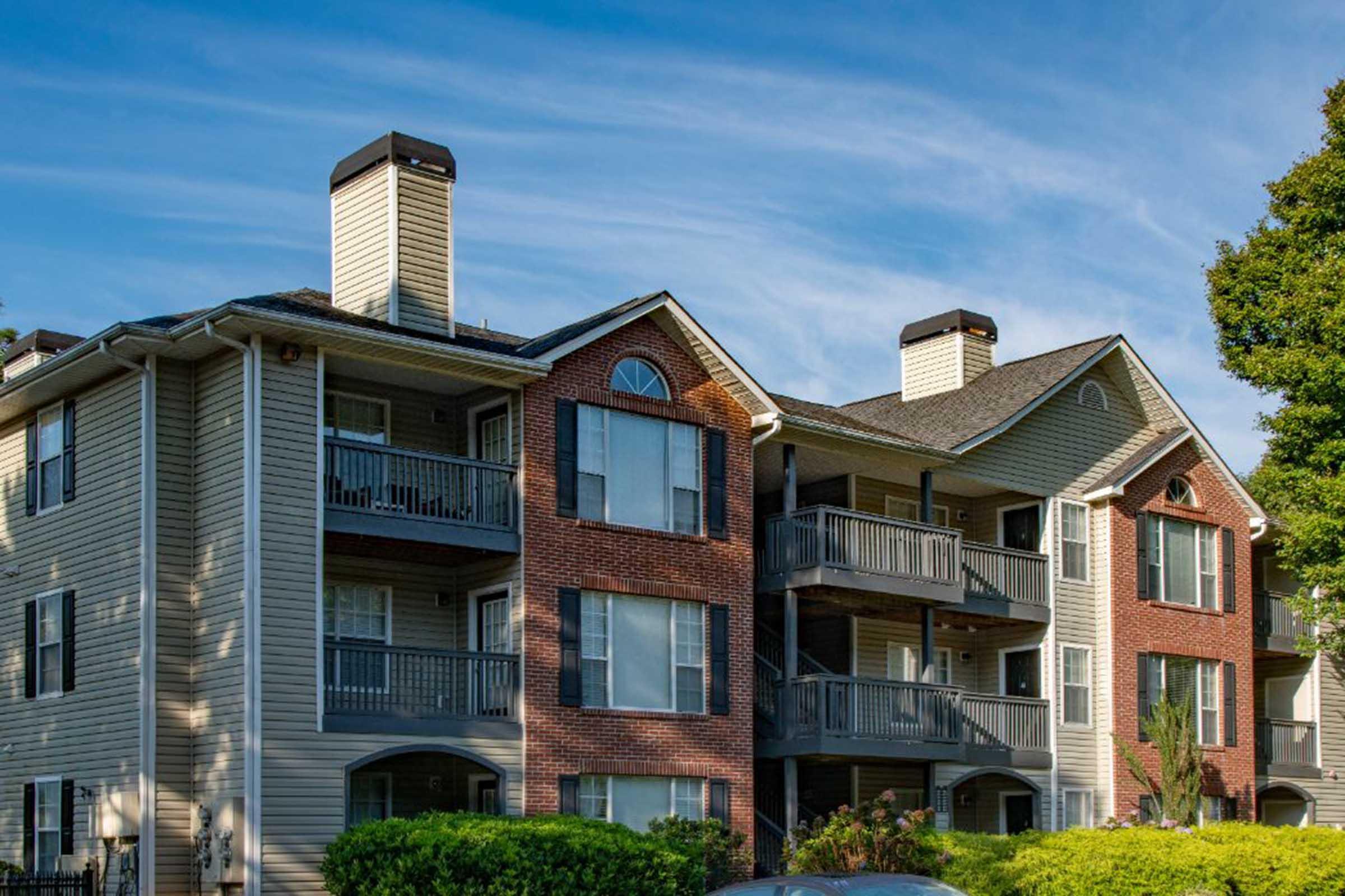 Two multi-story apartment buildings with brick and siding exteriors. The buildings feature balconies, multiple windows, and chimneys. The sky is clear and blue, and there is greenery around the base of the structures.