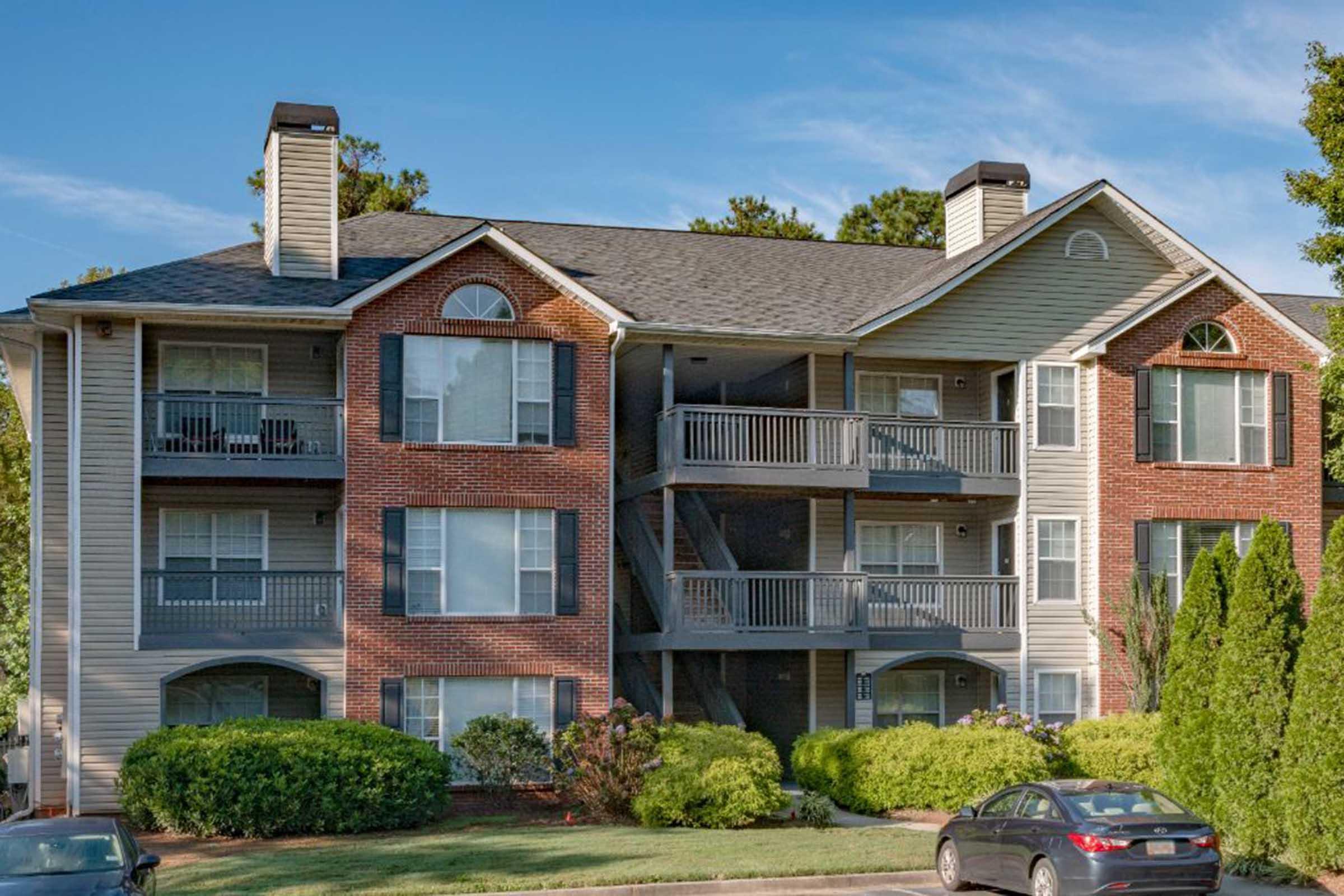 A multi-story apartment building featuring a mix of brick and siding exteriors, with balconies on each level. The landscape includes neatly trimmed shrubs and a few small trees, with a parked car in the foreground. Clear blue skies are visible above.