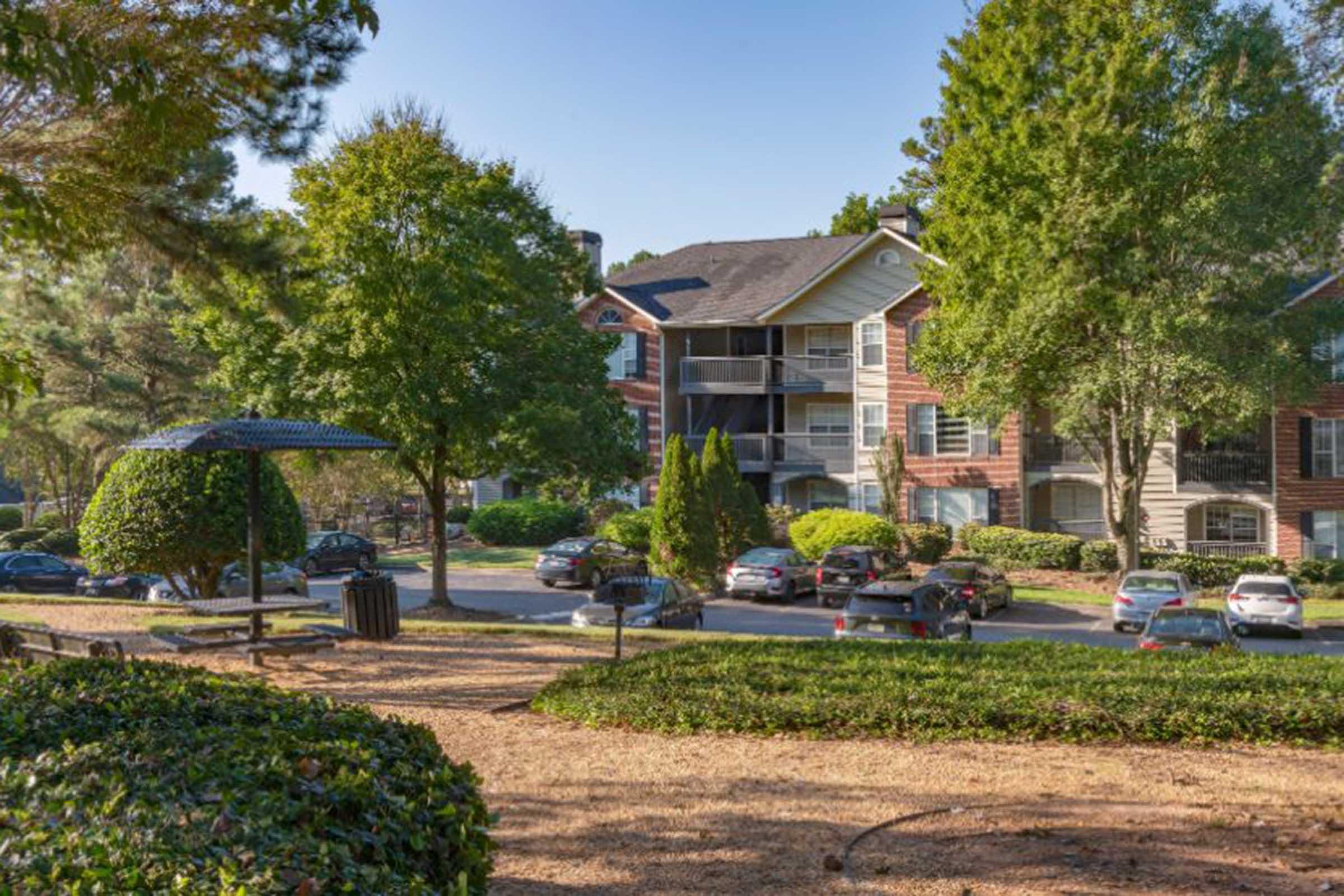 A residential apartment complex surrounded by trees and greenery. The foreground features a park-like area with a bench and a trash can. Several parked cars are visible in the driveway, and the buildings have balconies and brick facades, under a clear blue sky.