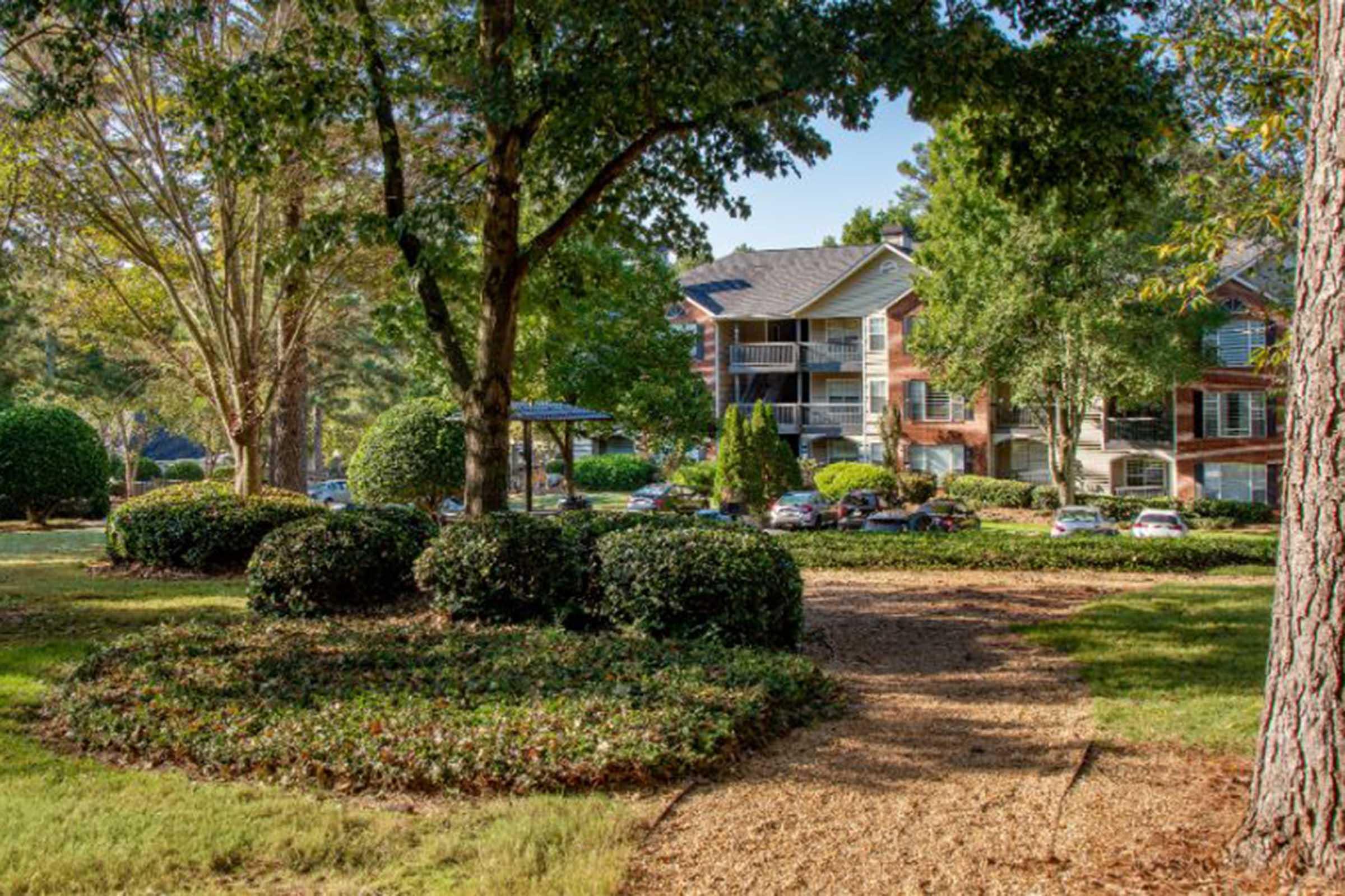 A landscaped pathway winds through a well-maintained green area featuring bushes and trees, leading to a cluster of apartment buildings in the background. The scene is bright and inviting, showcasing a peaceful outdoor setting with cars parked nearby and clear blue skies overhead.