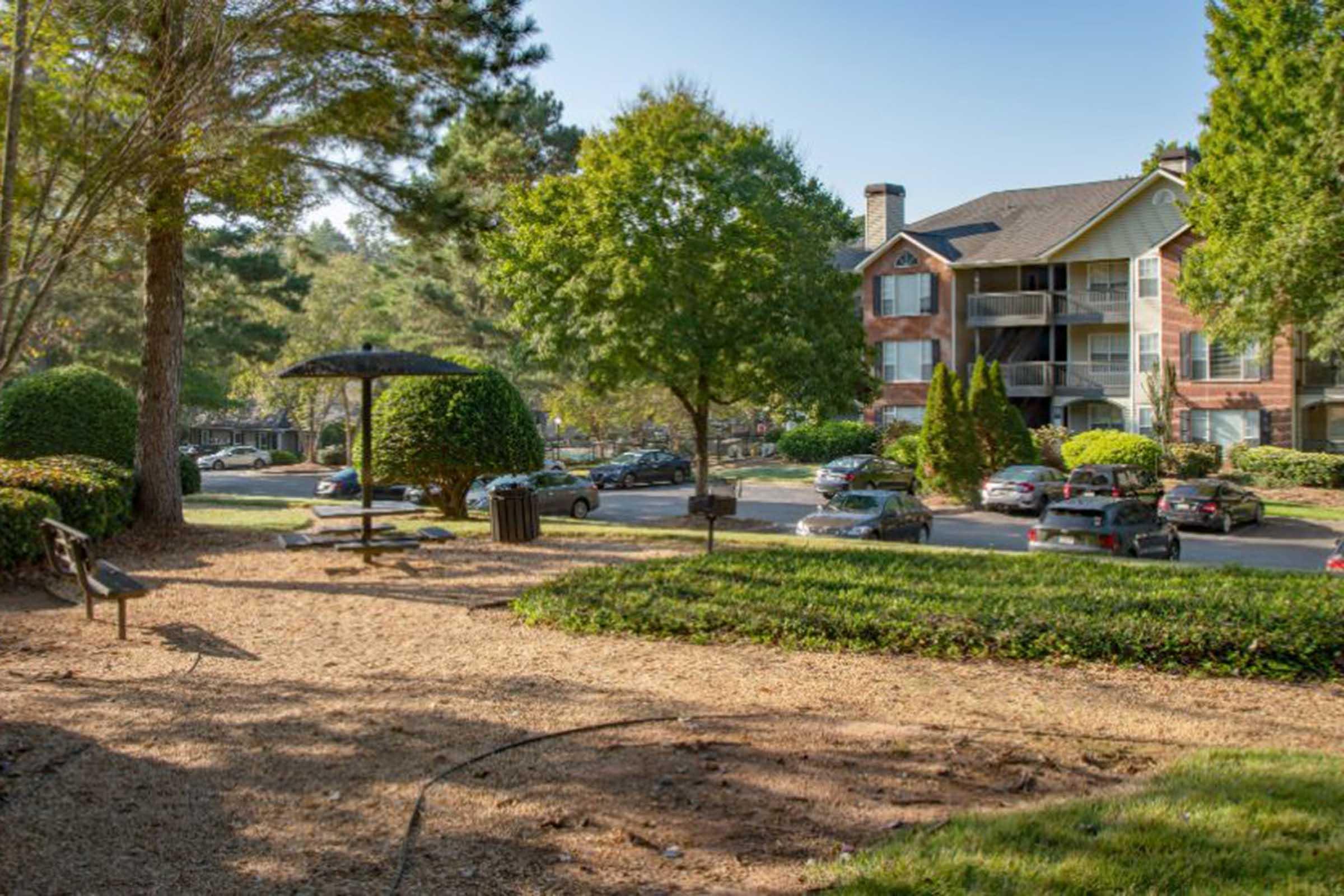 A view of a landscaped area with a bench and a sheltered seating space, surrounded by trees and shrubs. In the background, there are several parked cars and an apartment building with balconies. The scene is bright and inviting, showcasing a blend of greenery and residential life.