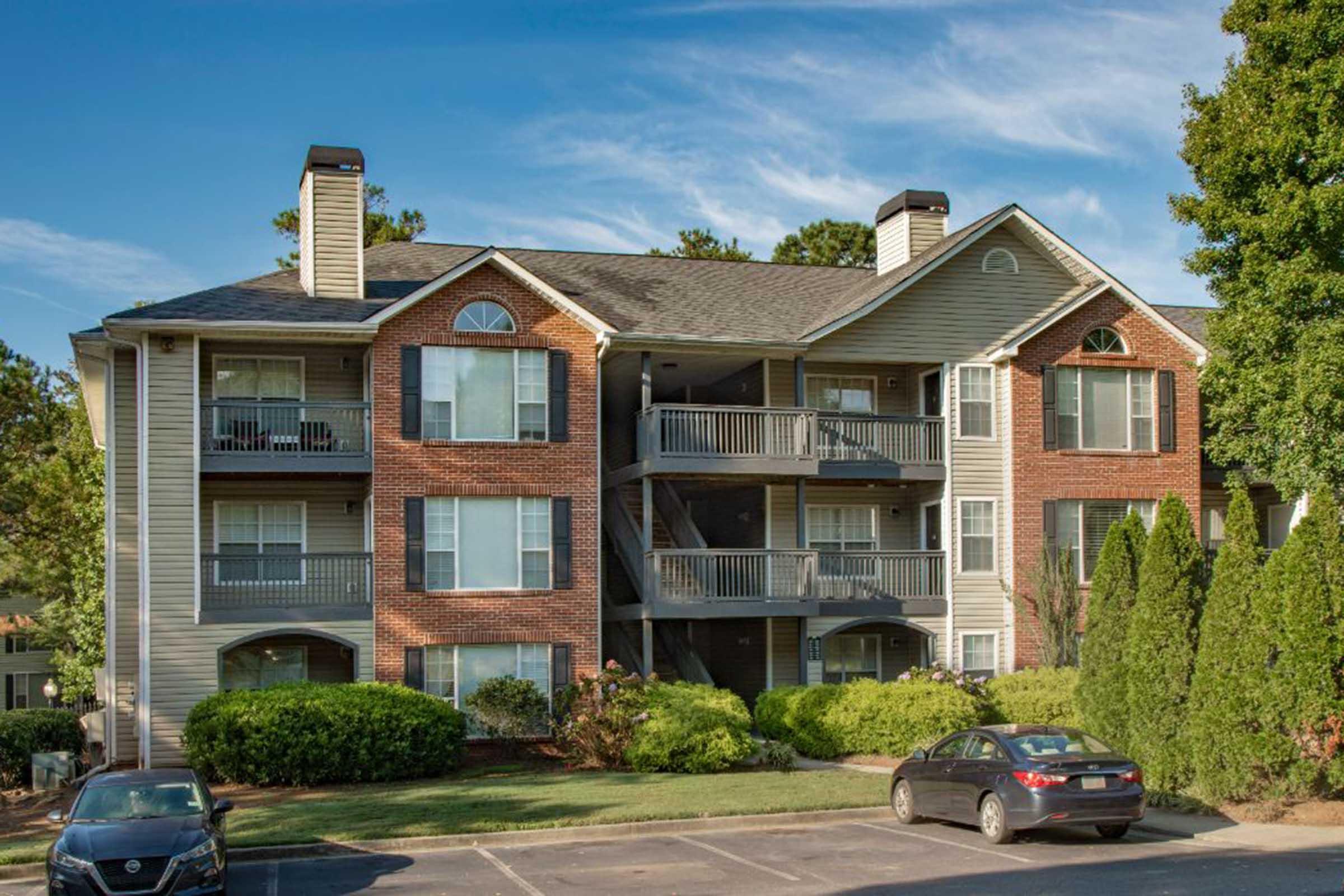 A three-story apartment building with a combination of brick and siding exterior. It features several balconies and large windows. Surrounding greenery includes shrubs and trees, and there is a parking lot in front with parked cars. The sky is clear and blue, indicating a sunny day.