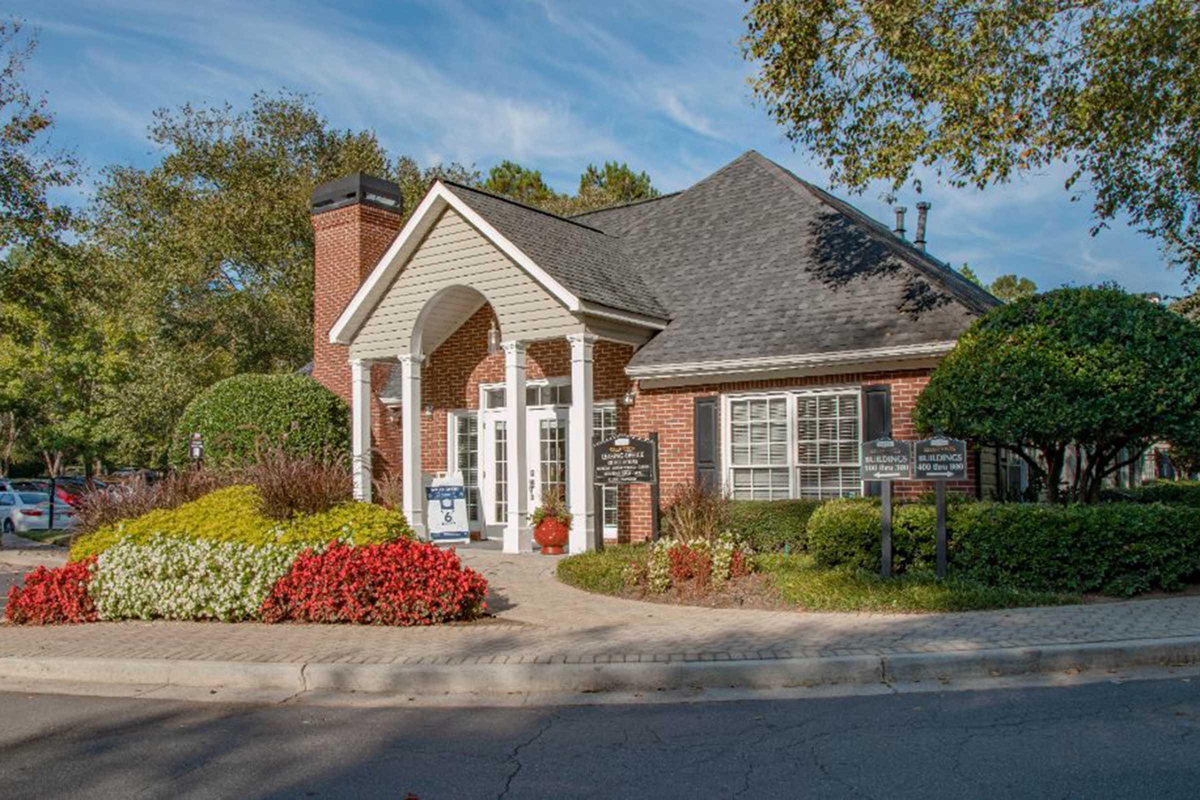 A beautifully landscaped building with brick architecture and a gabled roof, surrounded by colorful floral arrangements. The entrance features large windows and a double door, with signs to guide visitors. Lush greenery and trees are visible in the background, creating a welcoming atmosphere.