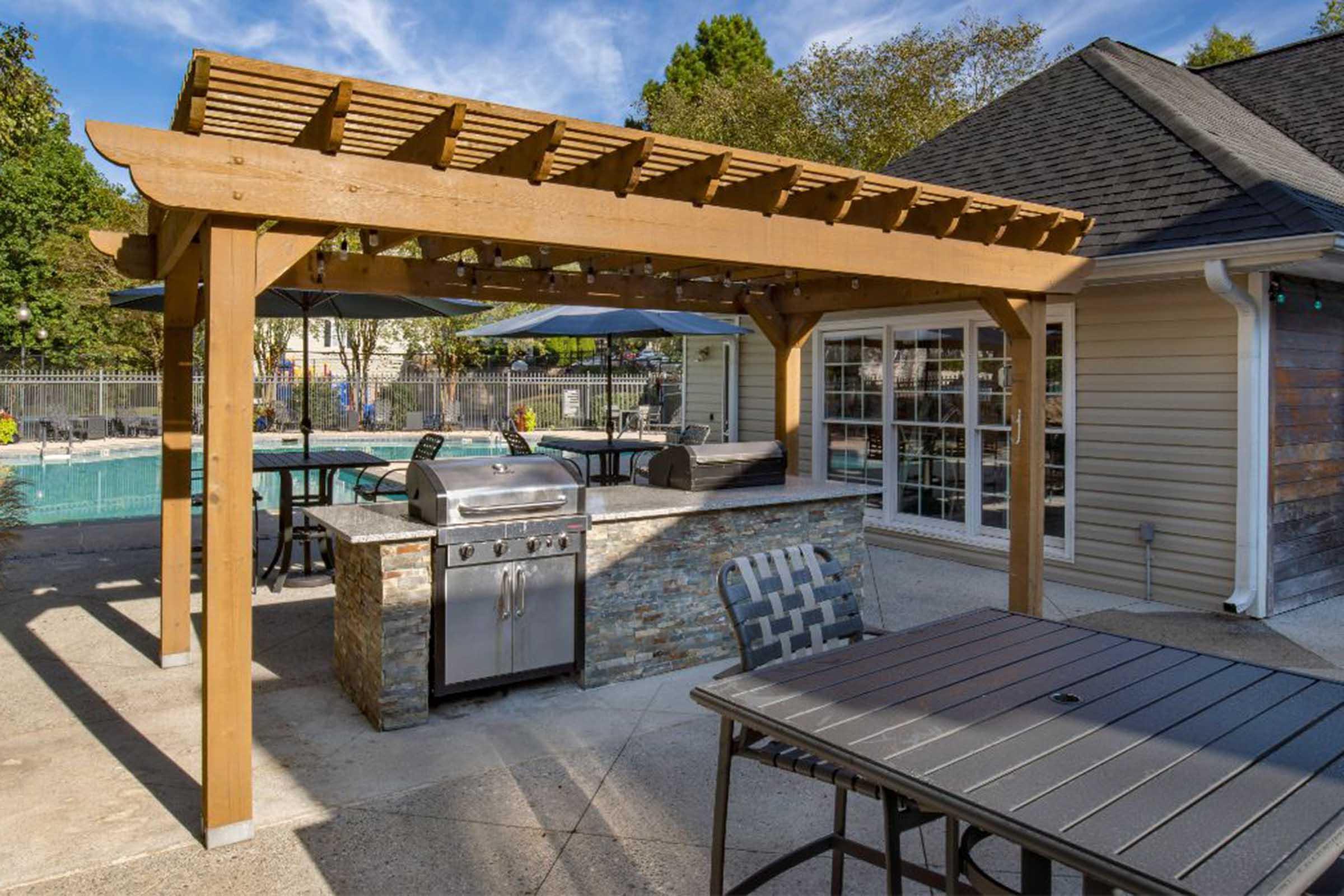 Outdoor area featuring a pergola over a patio with a built-in barbecue grill and stone base. Tables and chairs are arranged around a swimming pool in the background, creating a relaxed dining and entertainment space. Lush greenery and blue skies enhance the inviting atmosphere.
