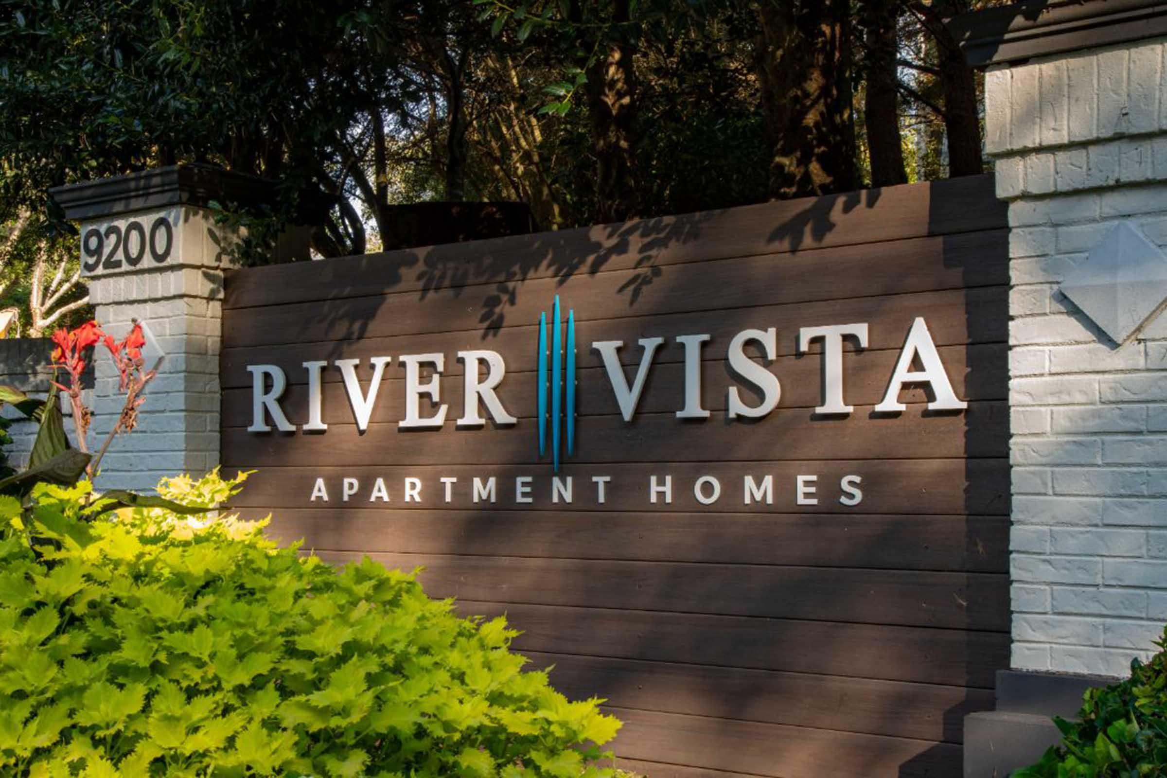 Sign for River Vista Apartment Homes, featuring bold lettering and decorative elements. The sign is set against a backdrop of greenery, with vibrant plants in the foreground. The address "9200" appears on one side of the sign, indicating the location of the apartments.