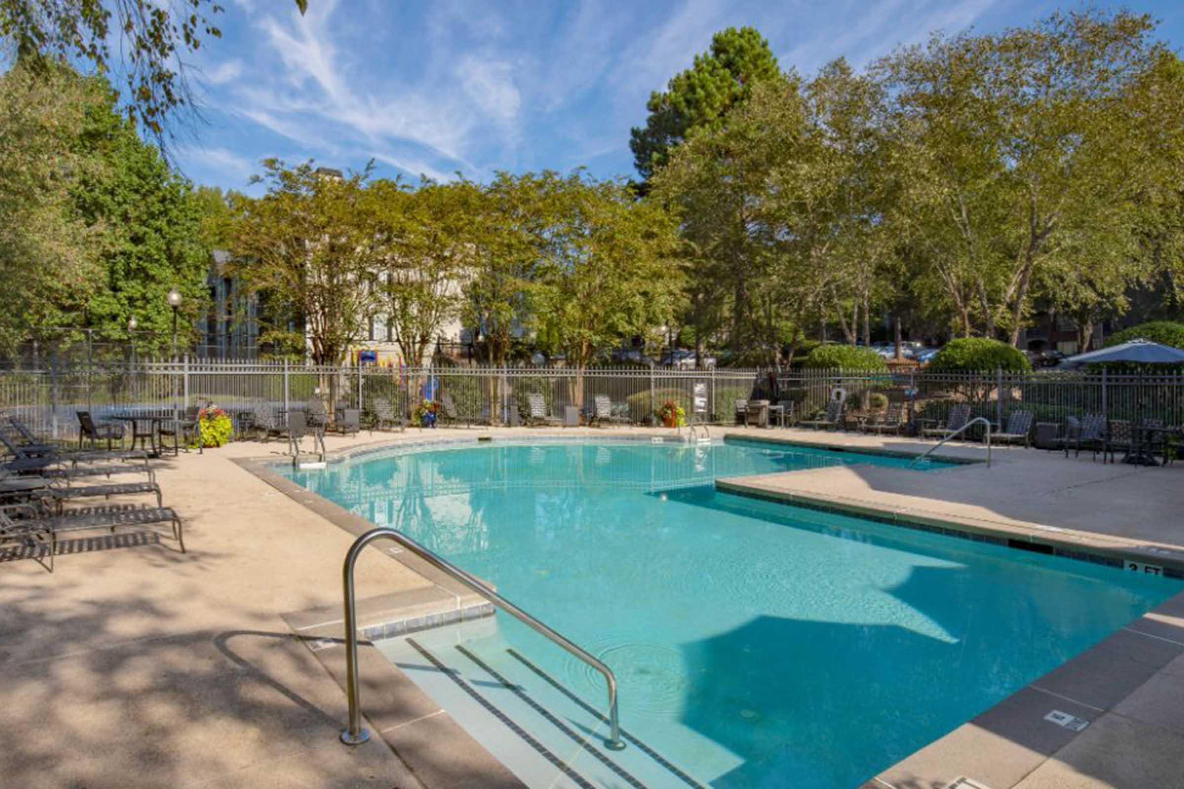 A serene outdoor swimming pool surrounded by lush green trees and lounge chairs. The pool features a clear blue water surface and a gradual slope entry. In the background, there are buildings and shaded sitting areas, creating a relaxing environment.