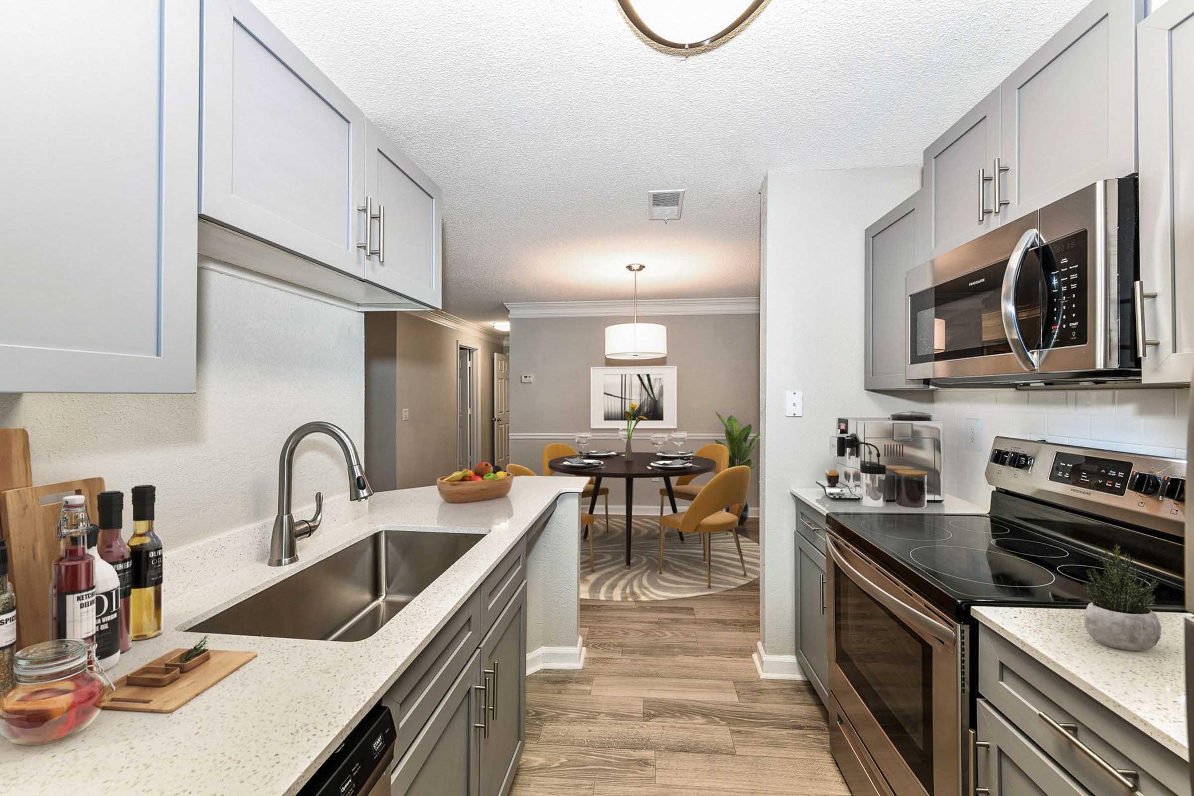 A modern kitchen featuring light gray cabinetry, a stainless steel sink, and a countertop with various kitchen items. The open layout leads to a dining area with a round table and yellow chairs, illuminated by a circular pendant light. The flooring is light wood, enhancing the contemporary feel.