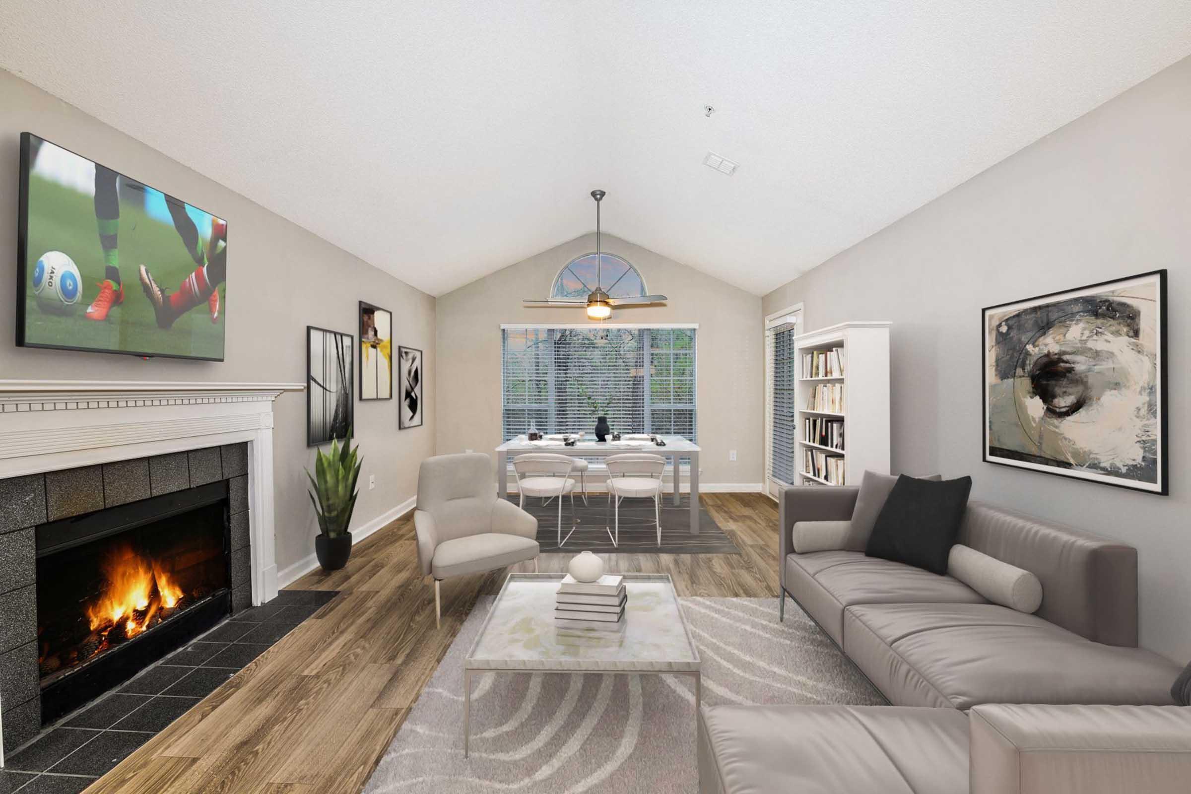 A modern living room featuring a cozy fireplace, a large TV mounted on the wall showing a sports game, and stylish furniture. The space is bright, with large windows allowing natural light, and includes a dining area visible in the background. Decor is minimalistic with art on the walls and a patterned area rug.