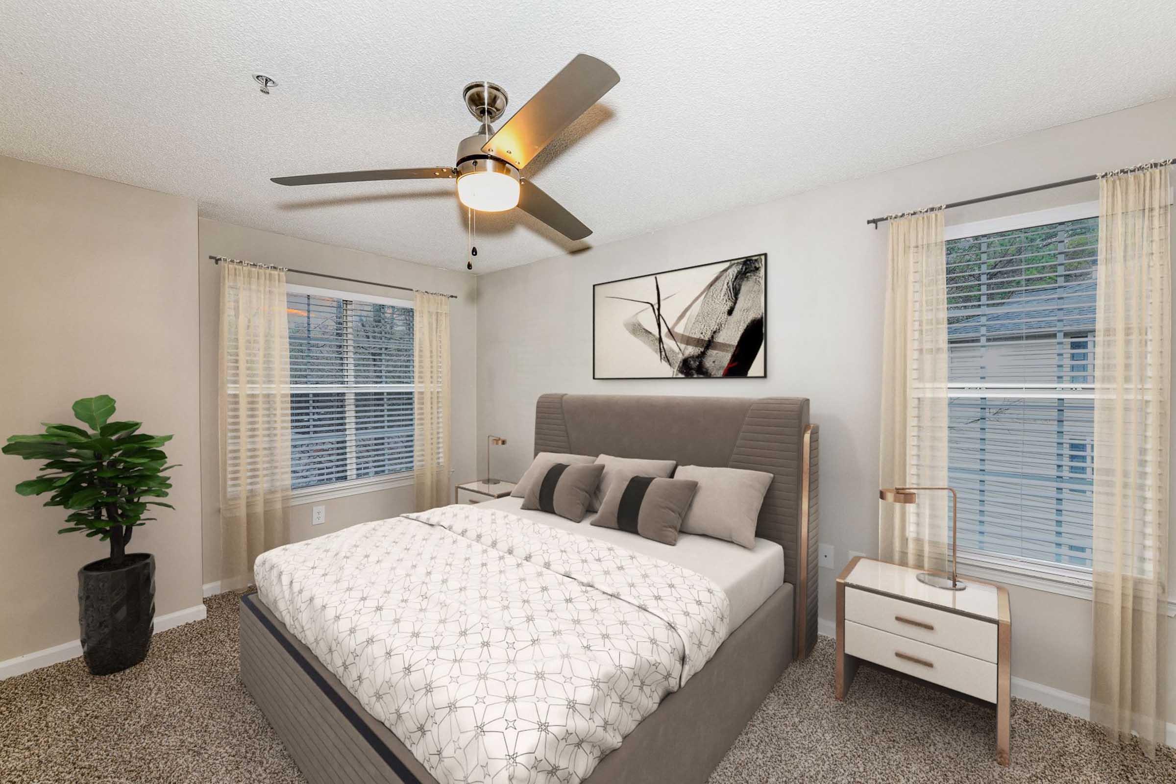 A cozy bedroom featuring a queen-sized bed with decorative pillows, a stylish nightstand, and a ceiling fan. Natural light streams through two windows with sheer curtains, and a decorative plant adds a touch of greenery to the space. A framed artwork is mounted on the wall above the bed.