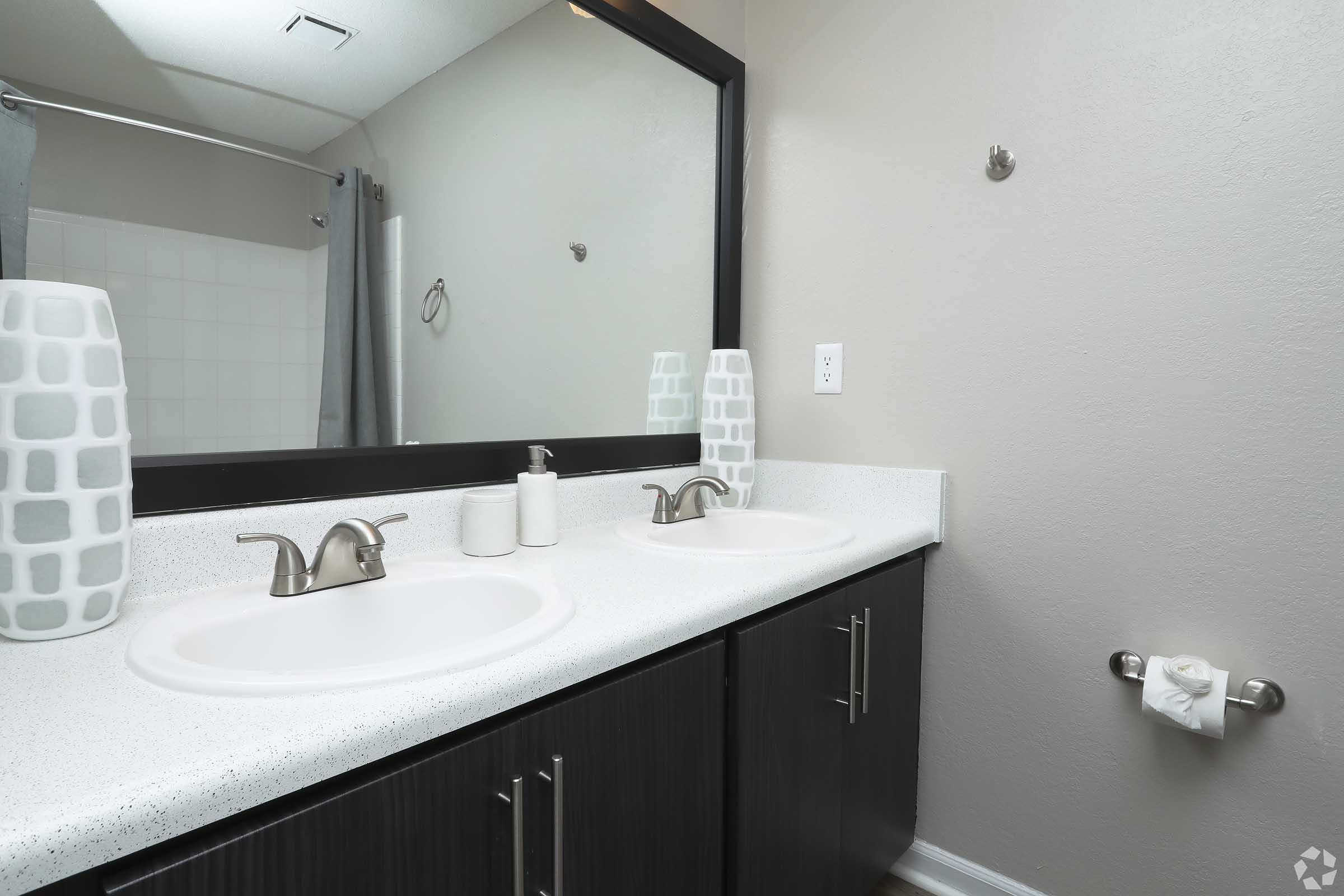 A modern bathroom featuring a double vanity with two white sinks, stylish faucets, and minimalist decor. The countertop is light-colored and smooth, with two decorative bottles. A large mirror spans the back wall, and there's a towel rack and toilet paper holder nearby. Soft gray curtains are drawn in the shower area.