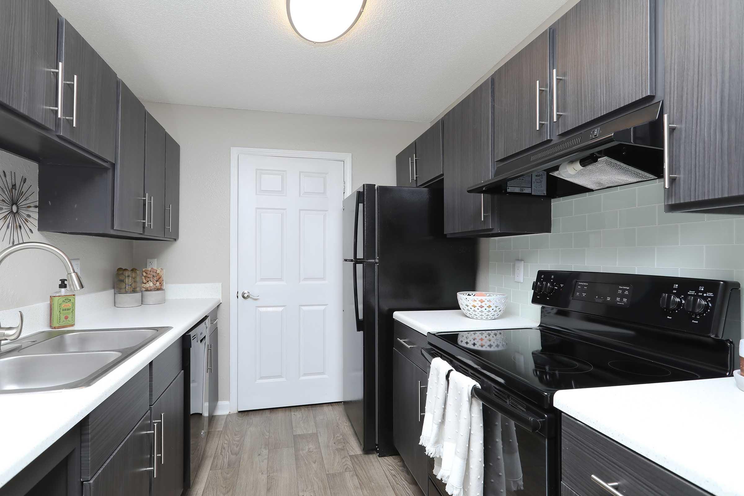 Modern kitchen featuring dark cabinetry with silver handles, a black refrigerator, and a black stove. The countertops are white, complemented by a light backsplash. A stainless steel sink is positioned under a light fixture. A white bowl decorates the countertop, adding a touch of style.