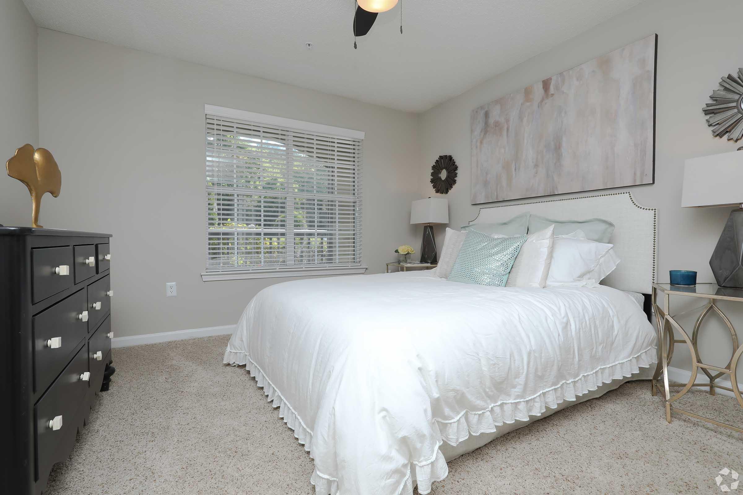 A cozy bedroom featuring a white bed with decorative pillows, a light-colored bedspread, and a plush carpet. There’s a black dresser to the left, two stylish bedside tables with lamps, and a large piece of abstract art hanging above the bed. Natural light flows in through a window with blinds.