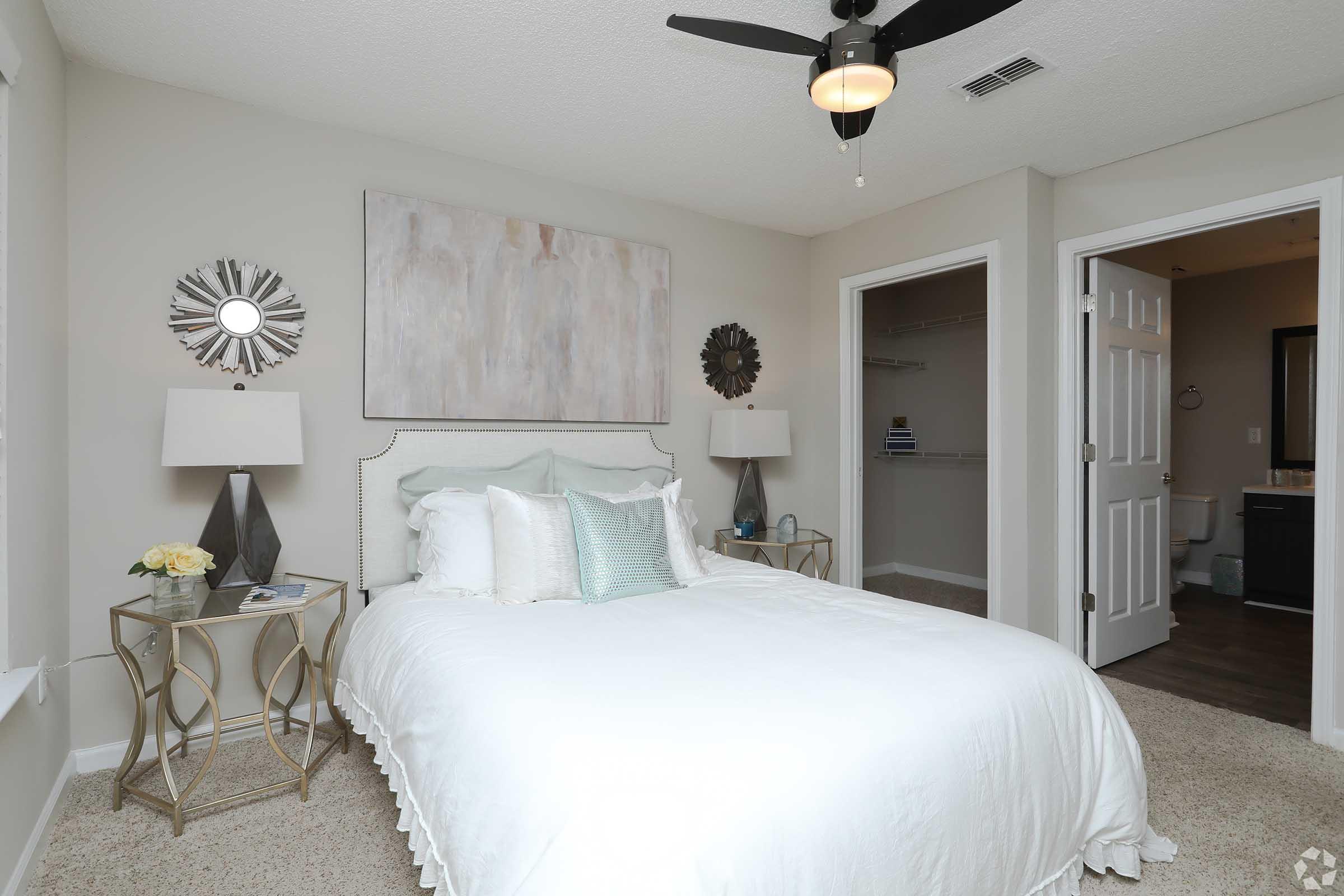 A cozy bedroom featuring a queen-sized bed with light bedding, decorative pillows, and a stylish headboard. The room has neutral walls, a ceiling fan, and two elegant lamps on either side of the bed. A mirrored wall decoration and a small nightstand add to the decor, along with an open closet and a bathroom visible through a door.